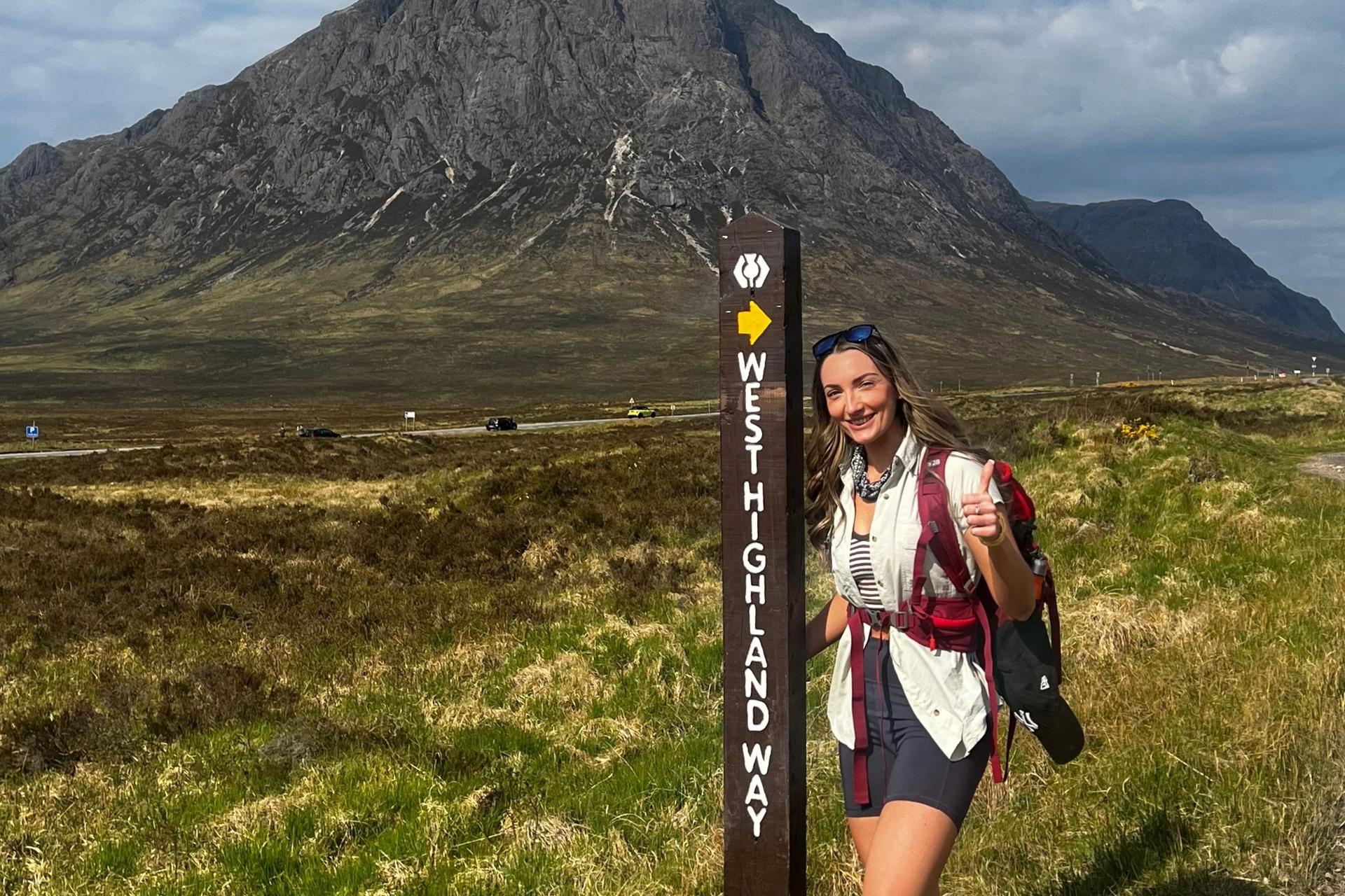 A hiker poses next to a West Highland Way signpost with a large mountain in the background