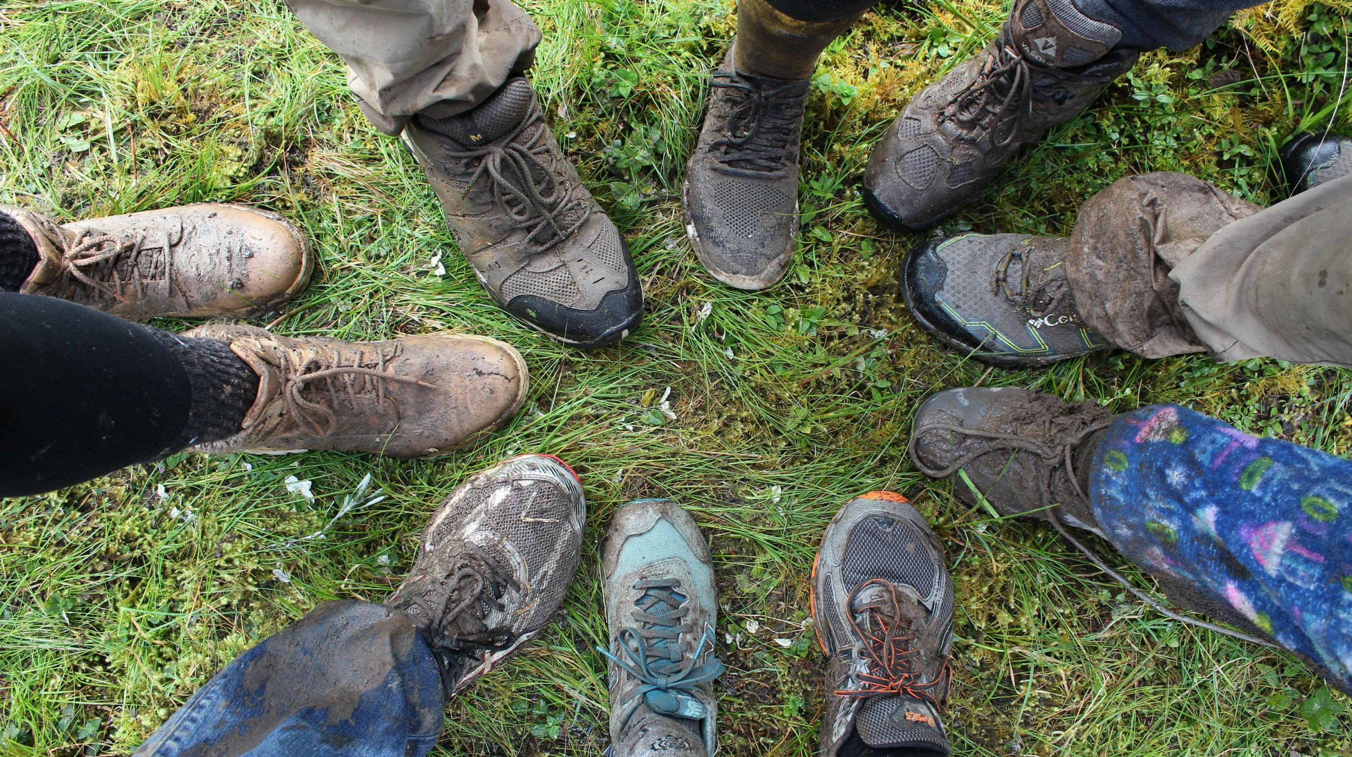 A top-down view of eight pairs of muddy shoes and boots standing in a circle on a grassy surface.