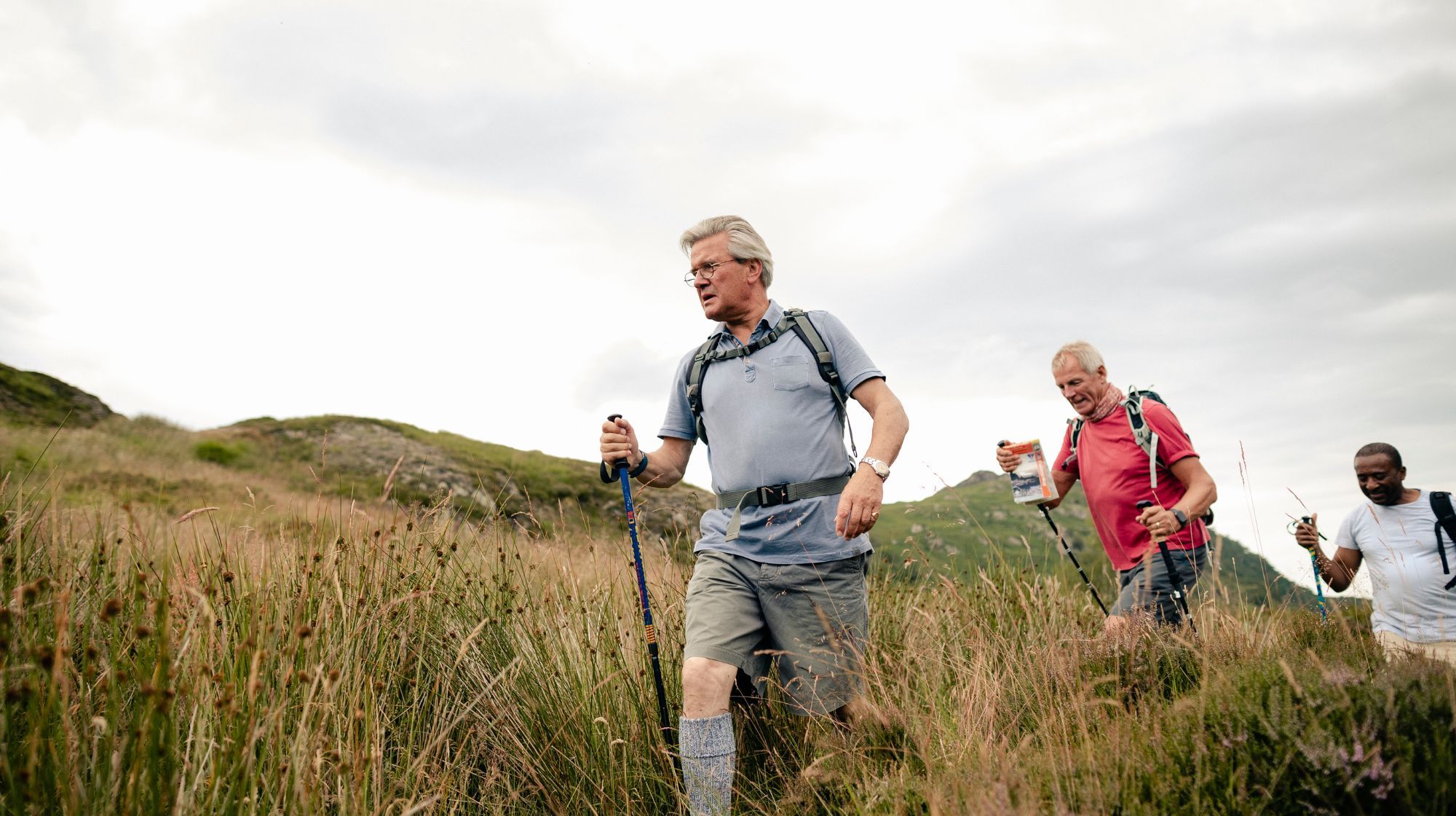 Three individuals hiking up a grassy hill with trekking poles under a cloudy sky.