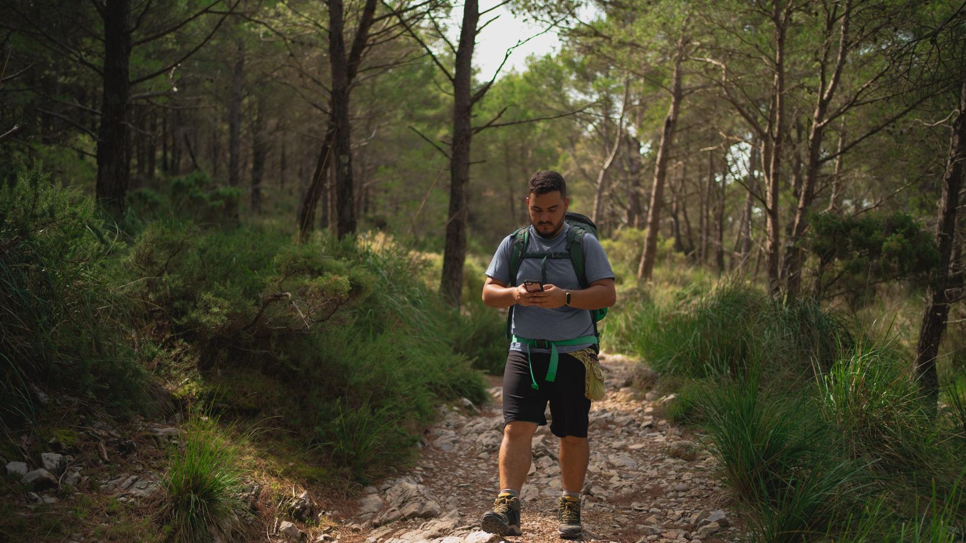 A person with a backpack is walking on a rocky path in a forest and looking at their phone.