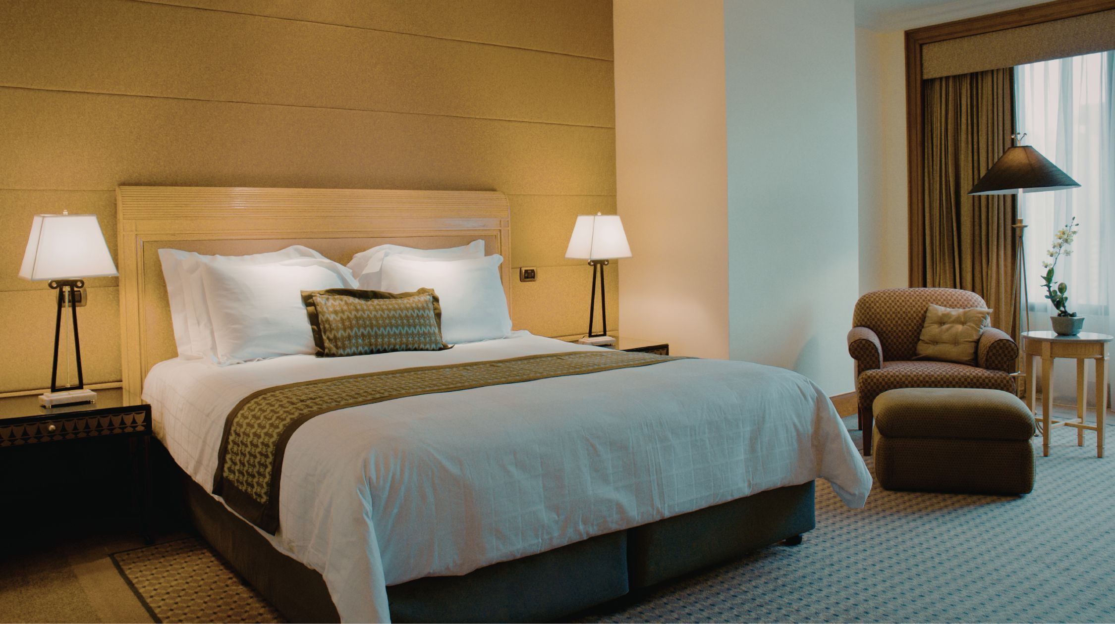 A well-made hotel bed with white linens and decorative pillows is shown next to a nightstand with a lamp, and a patterned armchair with an ottoman is visible near a window with curtains.