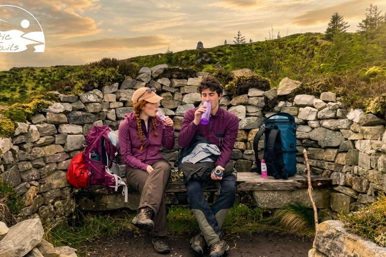 Two walkers taking a food and water break beside a dry-stone wall on a walking trail