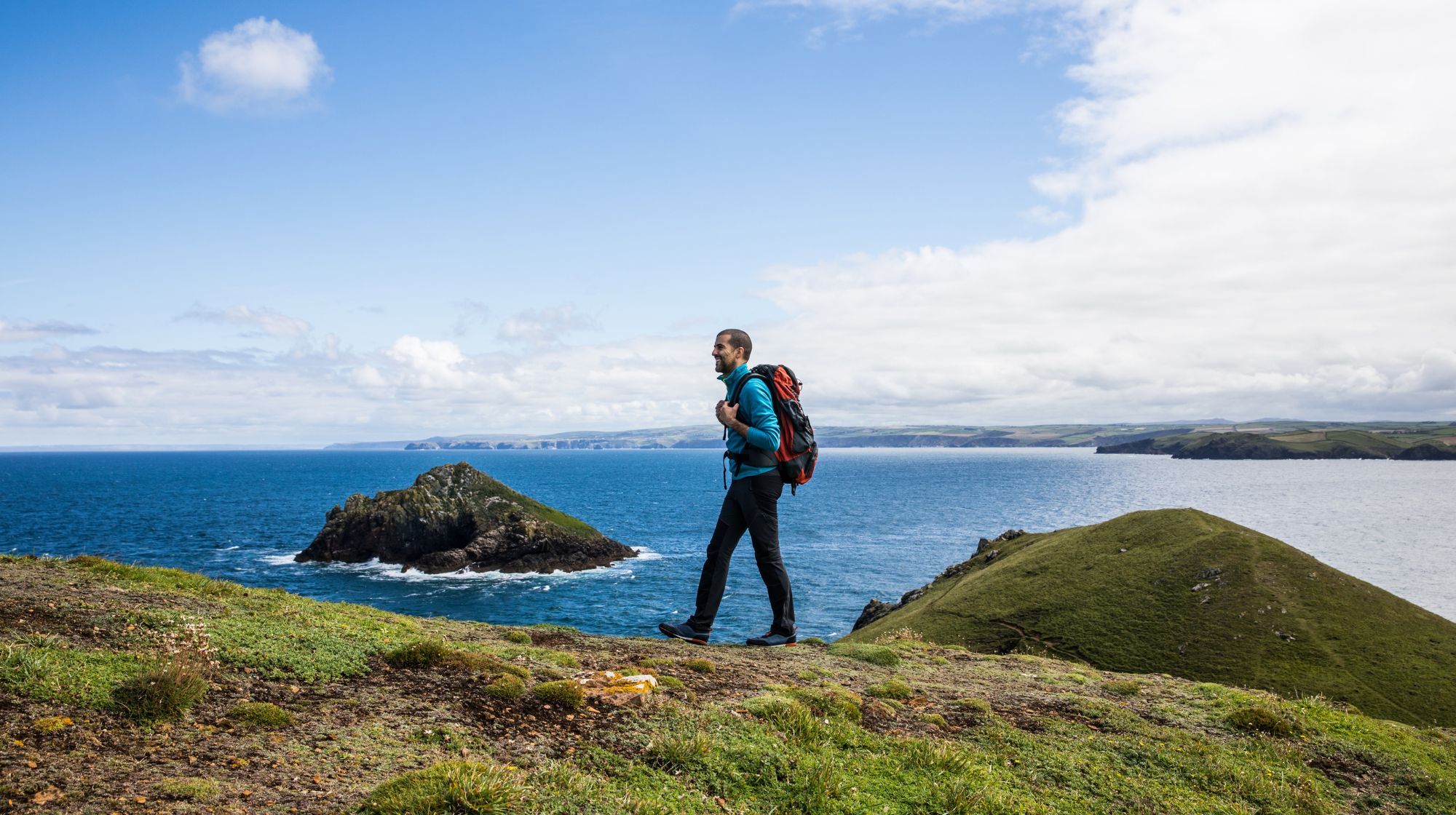 A person with a backpack walks along a grassy cliff overlooking the blue ocean under a cloudy sky.