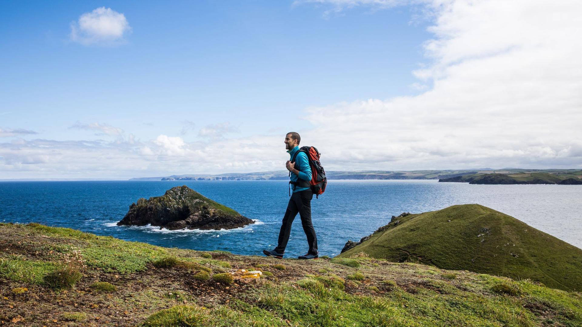 A person with a backpack walks along a grassy cliff overlooking the blue ocean under a cloudy sky.