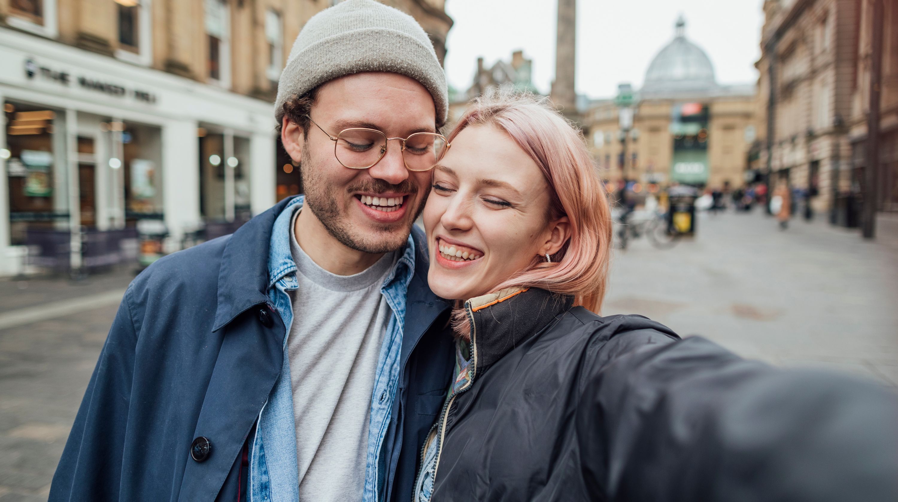 A couple taking a selfie on a city street, both smiling widely with their arms around each other. The background shows buildings and blurred figures.