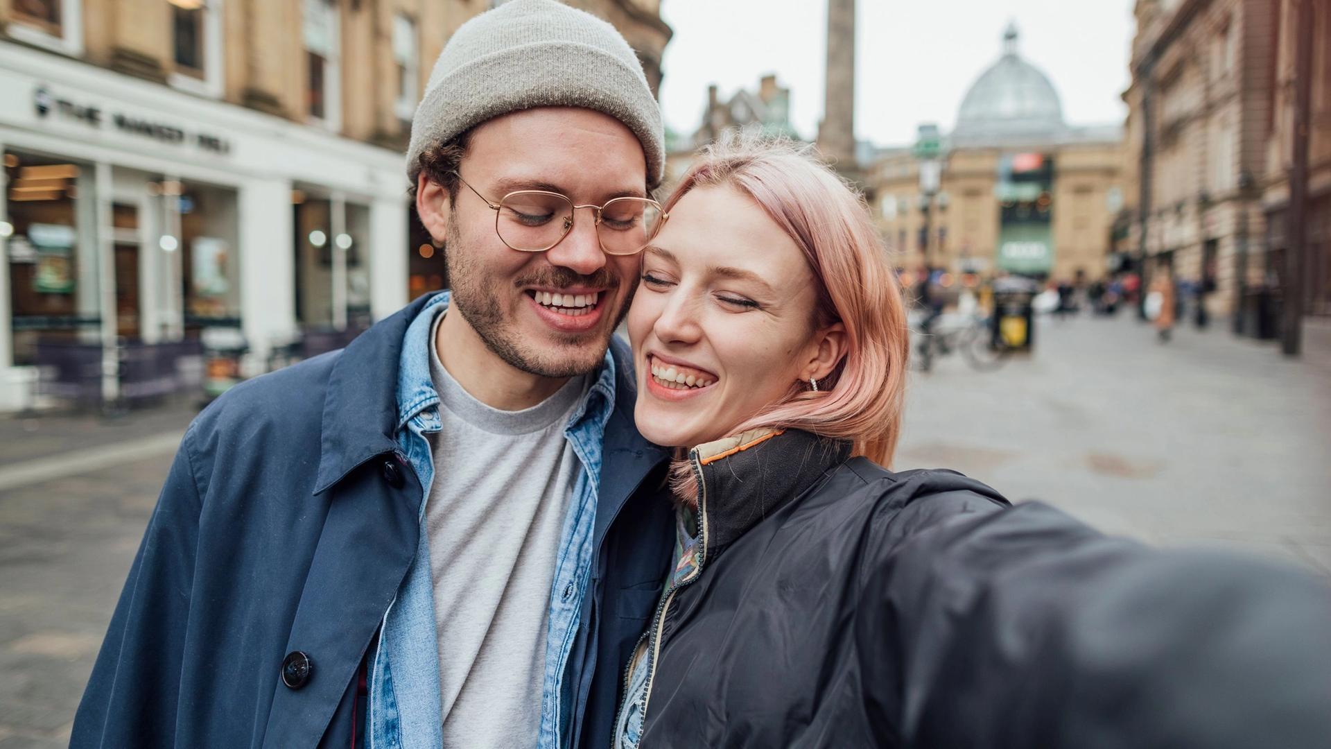 A couple taking a selfie on a city street, both smiling widely with their arms around each other. The background shows buildings and blurred figures.