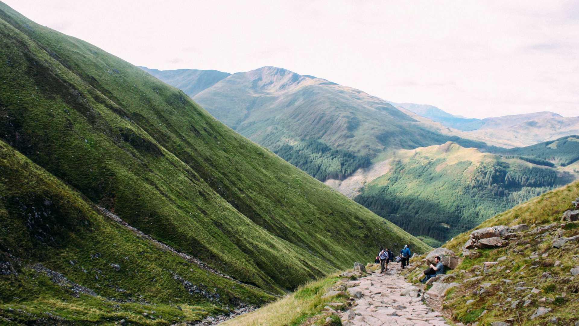 A group of hikers on a rocky path surrounded by lush green mountains under a hazy sky.