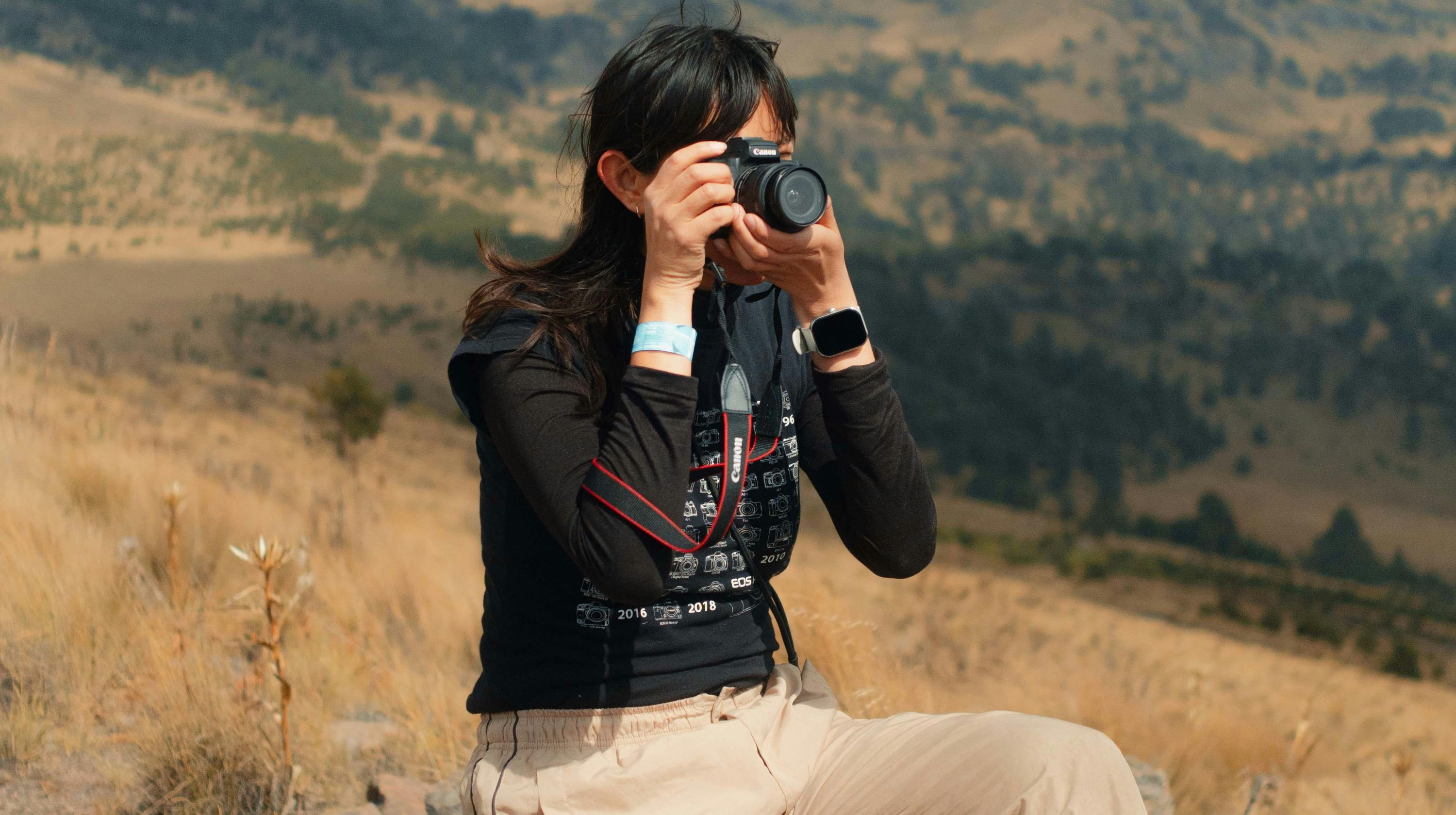 A person holds a camera up to their face, taking a photo in a dry, hilly landscape.