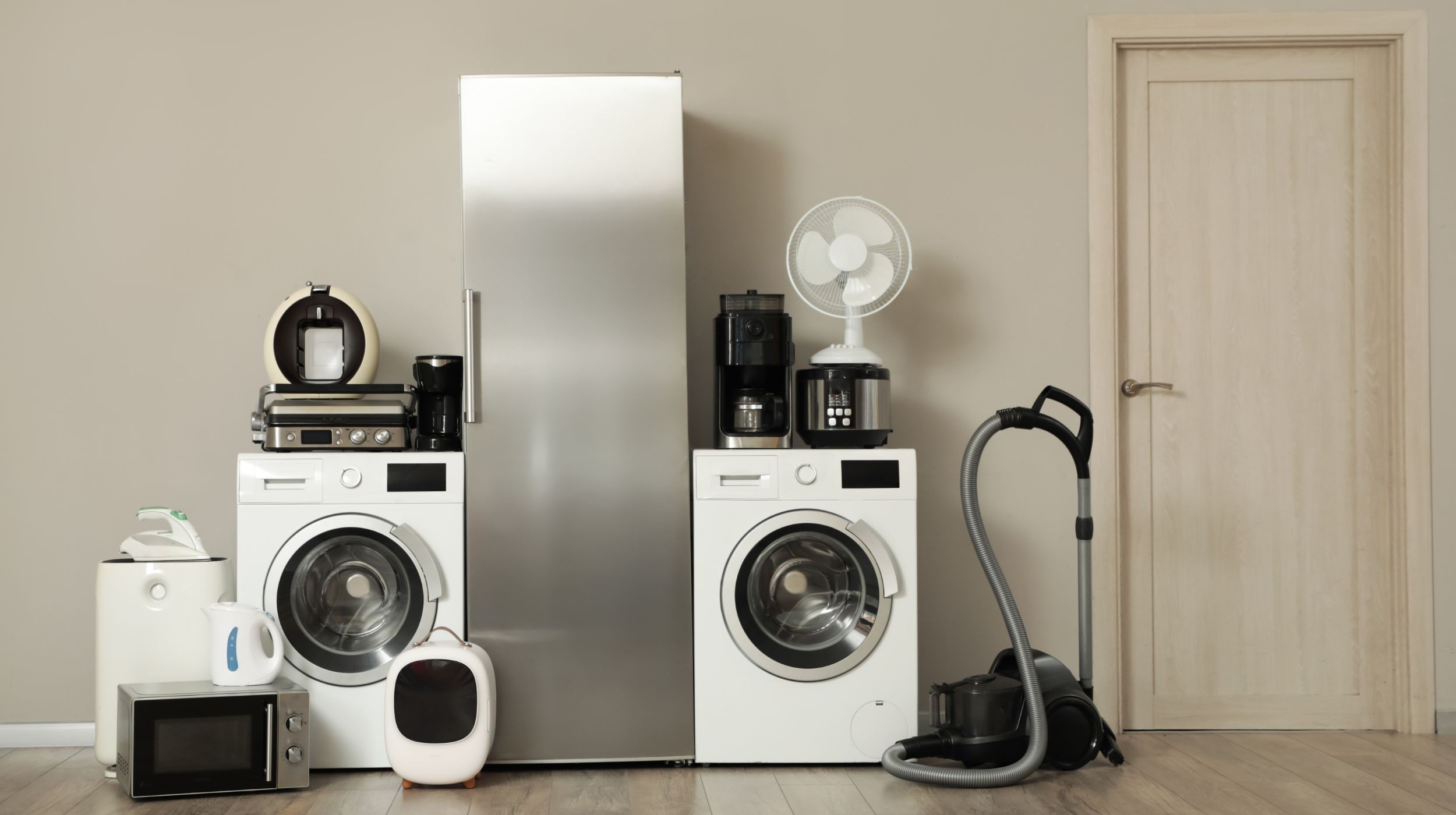 A collection of home appliances including a refrigerator, washing machines, microwave, coffee makers, fan, vacuum cleaner, and other kitchen gadgets are arranged on a wooden floor against a neutral wall with a closed door on the right.