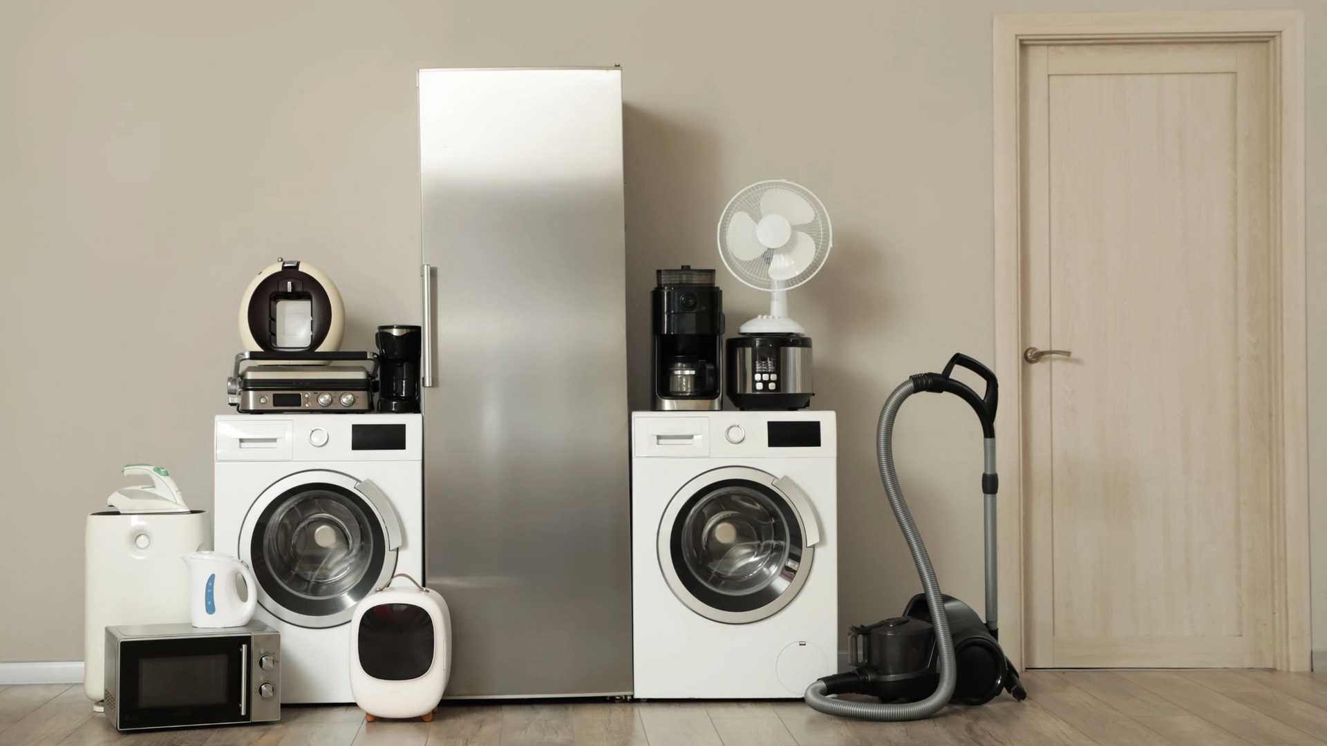 A collection of home appliances including a refrigerator, washing machines, microwave, coffee makers, fan, vacuum cleaner, and other kitchen gadgets are arranged on a wooden floor against a neutral wall with a closed door on the right.