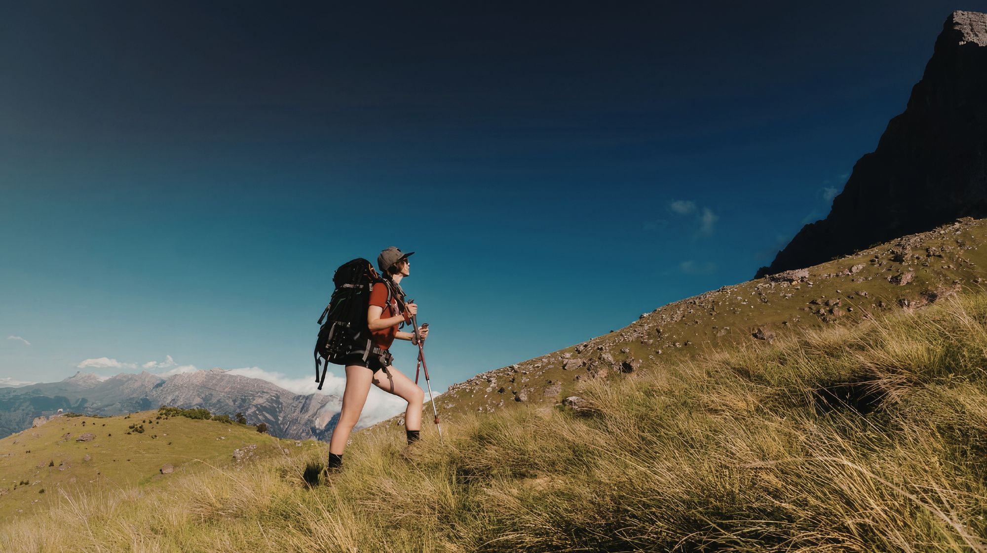 A hiker with a large backpack and trekking poles ascends a grassy mountain slope under a clear blue sky.