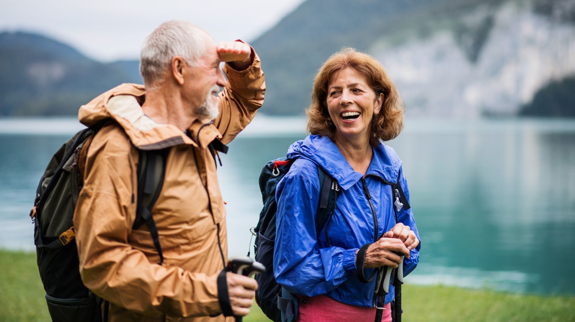 Two hikers with backpacks and trekking poles smile near a lake with mountains in the background. The person on the left shields their eyes from the sun.