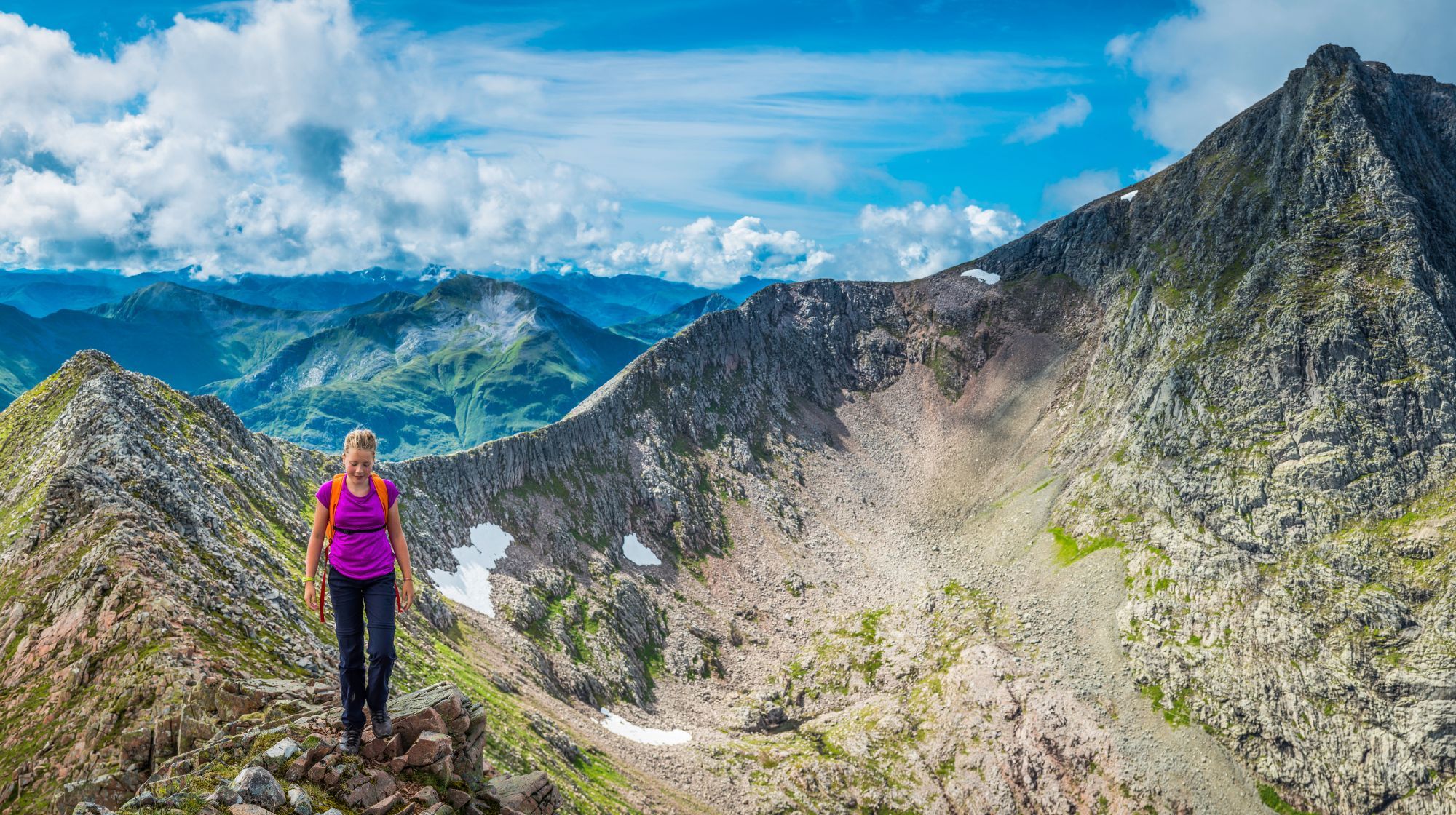 A person wearing a purple shirt and backpack hikes on a rocky mountain ridge with a vast mountain range in the background under a blue sky with clouds.