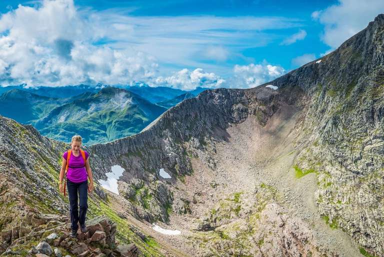 A person wearing a purple shirt and backpack hikes on a rocky mountain ridge with a vast mountain range in the background under a blue sky with clouds.