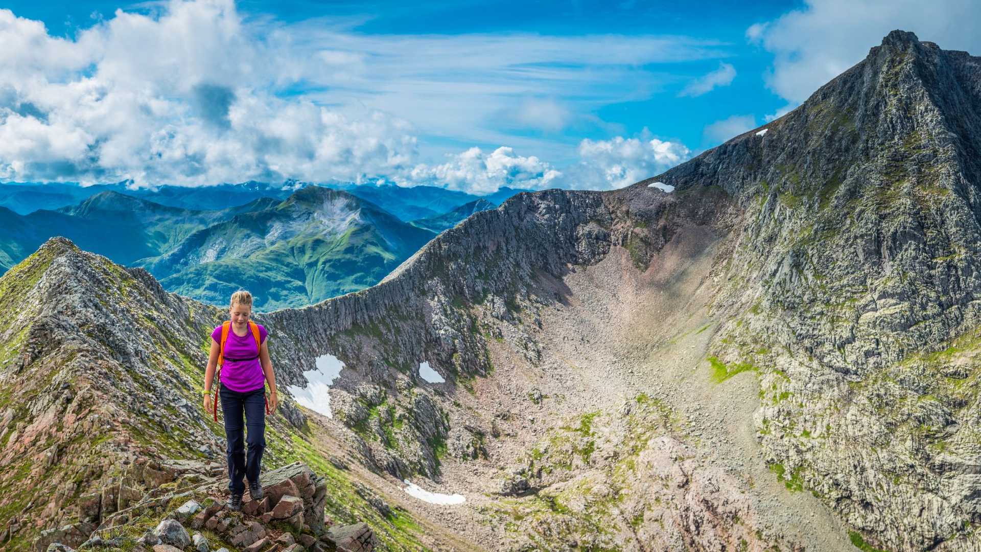 A person wearing a purple shirt and backpack hikes on a rocky mountain ridge with a vast mountain range in the background under a blue sky with clouds.