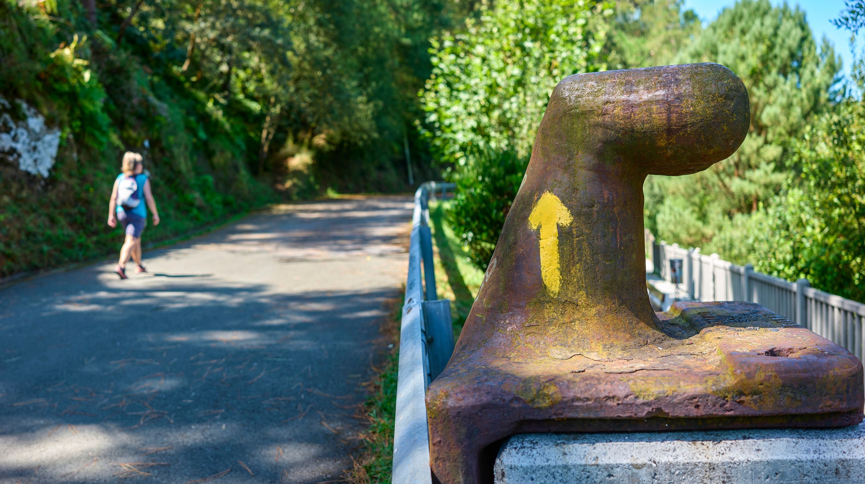 A weathered bollard with a yellow arrow pointing upwards stands beside a paved path where a person is walking away from the camera.