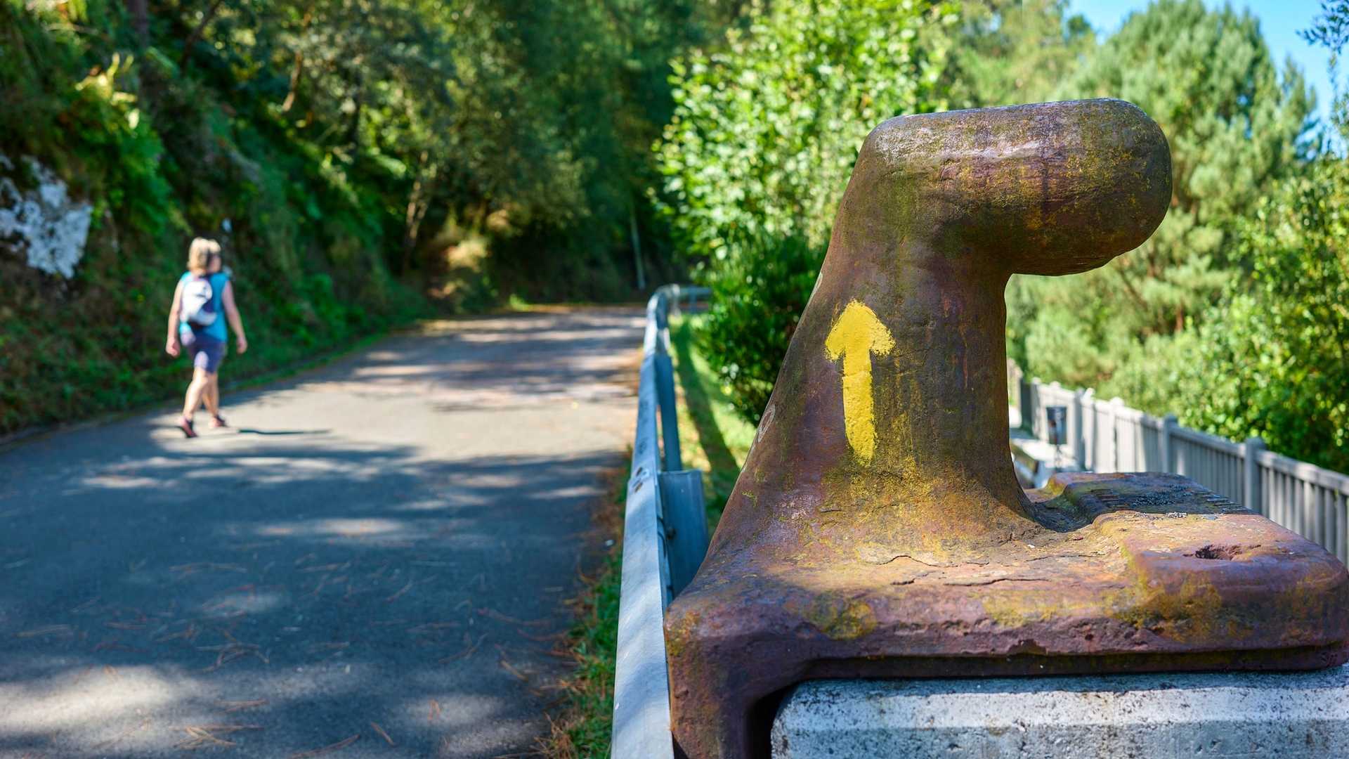 A weathered bollard with a yellow arrow pointing upwards stands beside a paved path where a person is walking away from the camera.