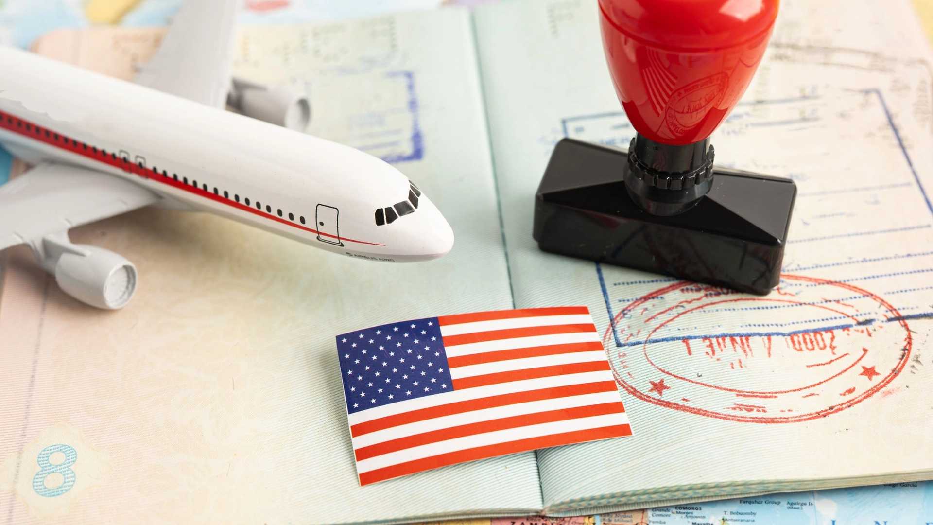 A toy airplane, a small American flag, and a red rubber stamp rest on an open passport with visa stamps.