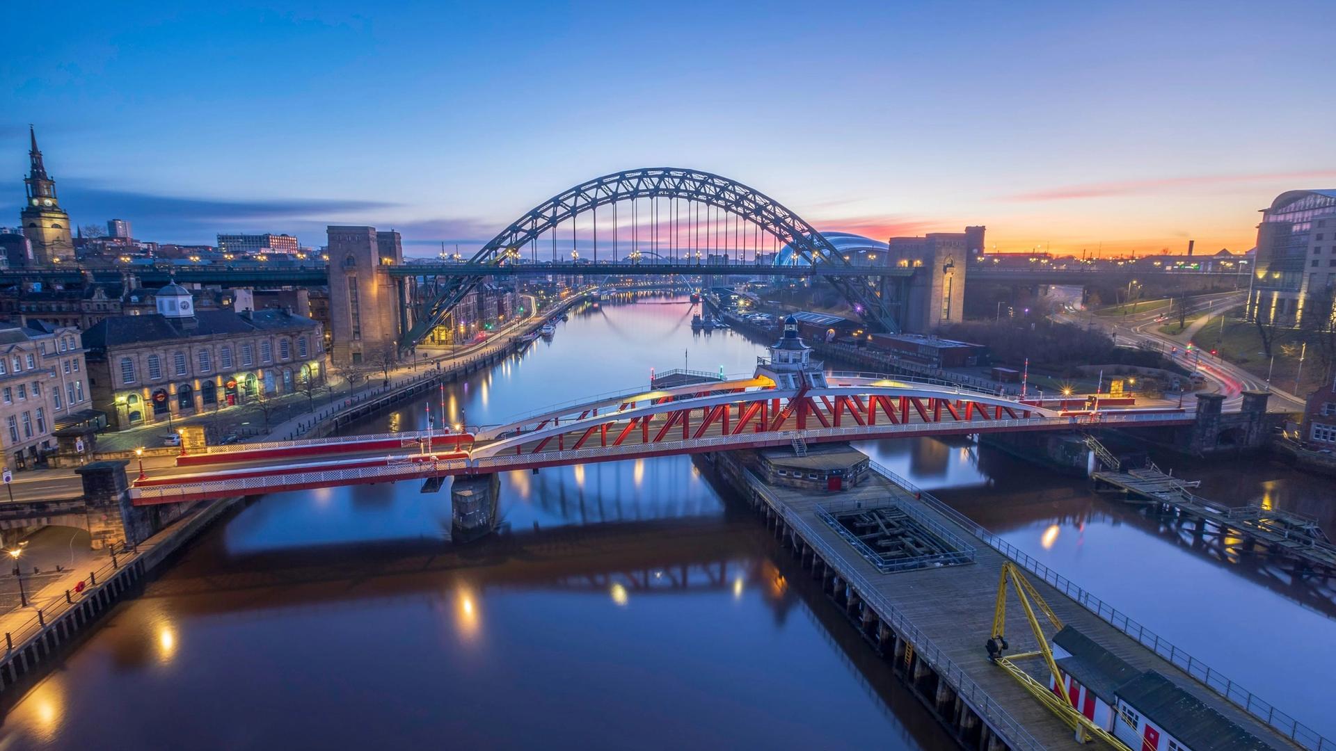 The Tyne Bridge and Swing Bridge span across the River Tyne at dusk, with city lights reflecting on the water and a colorful sky above.