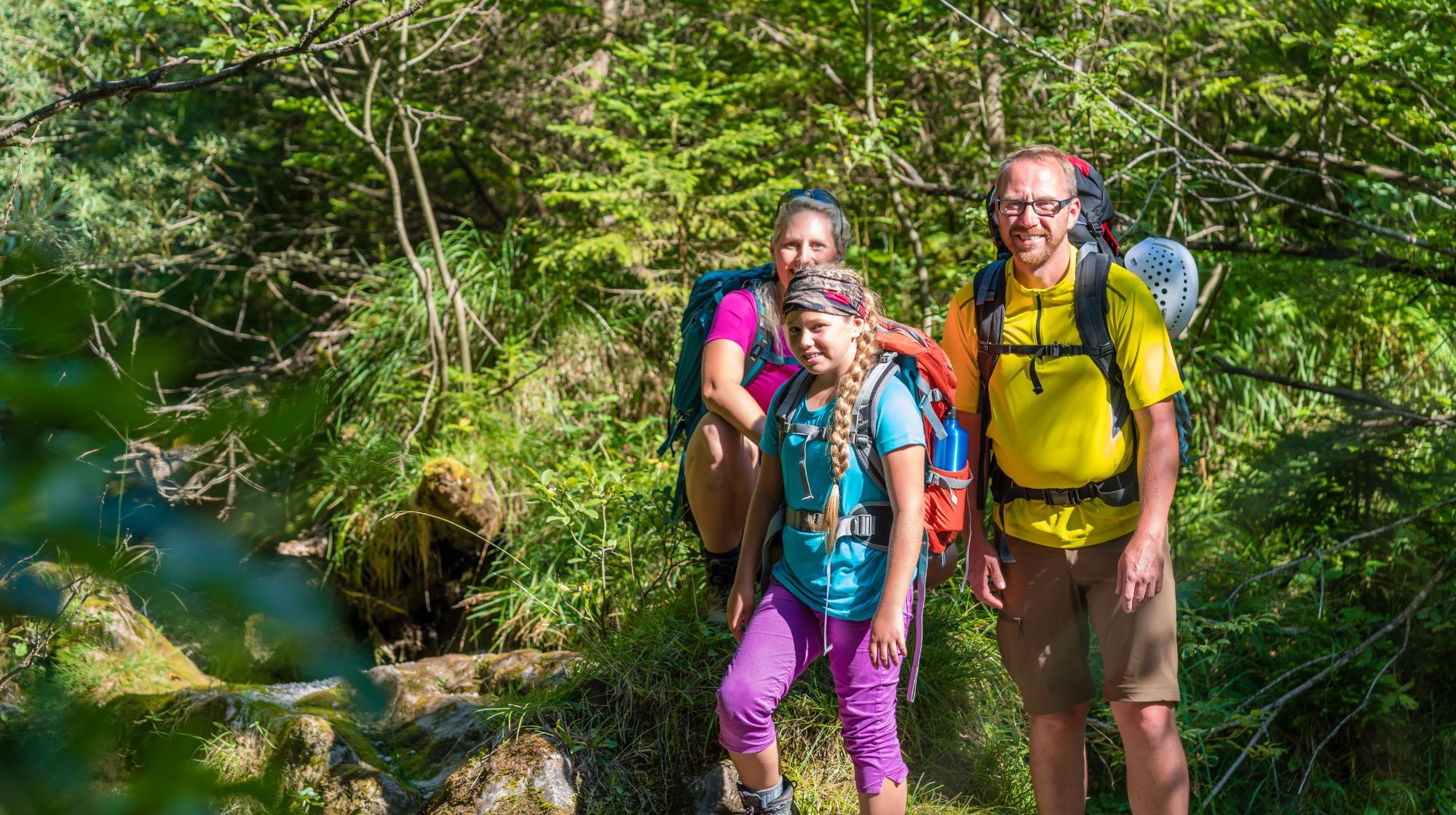 A family wearing backpacks hikes through a lush green forest with sunlight filtering through the trees.