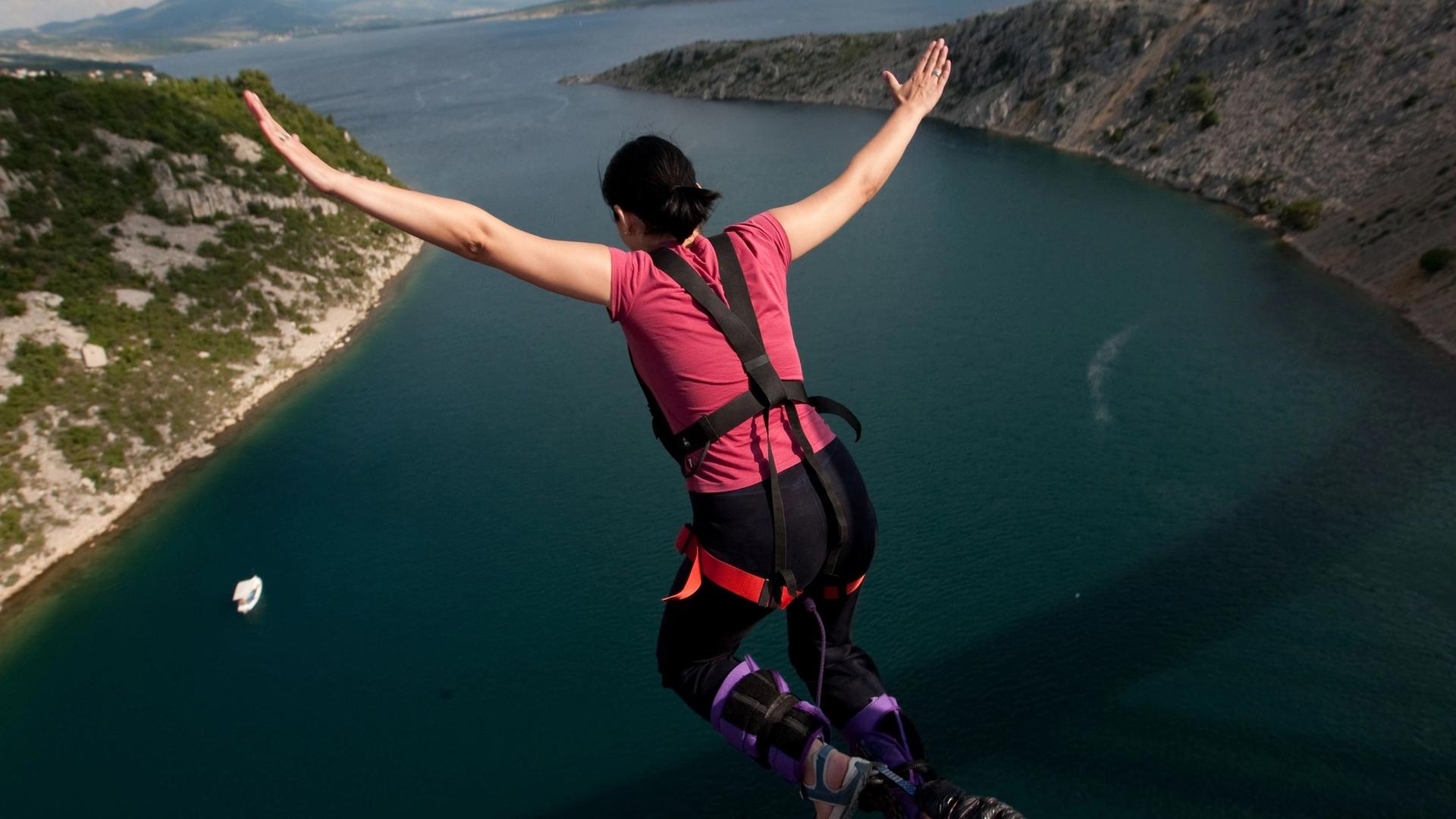 A person is bungee jumping over a deep blue body of water with rocky, green hillsides on either side.
