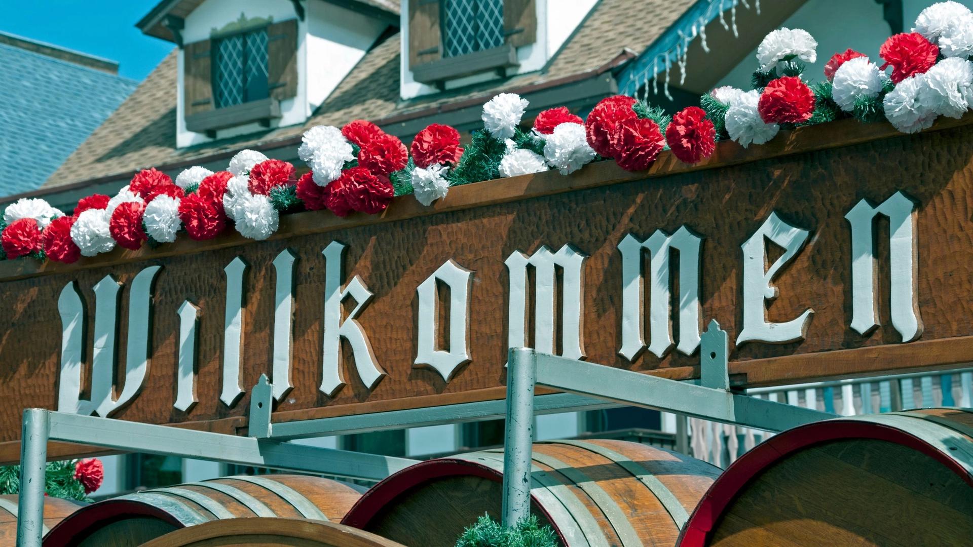 A wooden sign with the word "Willkommen" in white gothic lettering, decorated with red and white flowers. Below the sign are wooden barrels.