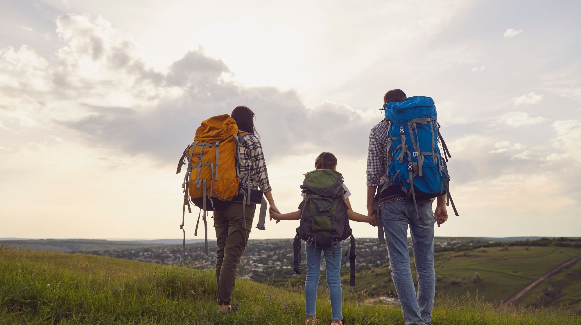 Family with large backpacks stand on a grassy hill, holding hands and looking out over a distant town under a cloudy sky.