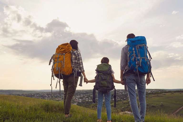 Family with large backpacks stand on a grassy hill, holding hands and looking out over a distant town under a cloudy sky.
