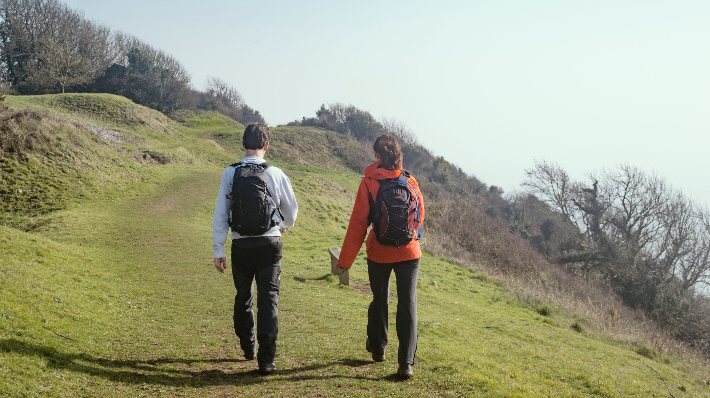 Two people with backpacks walk up a grassy hill on a sunny day.