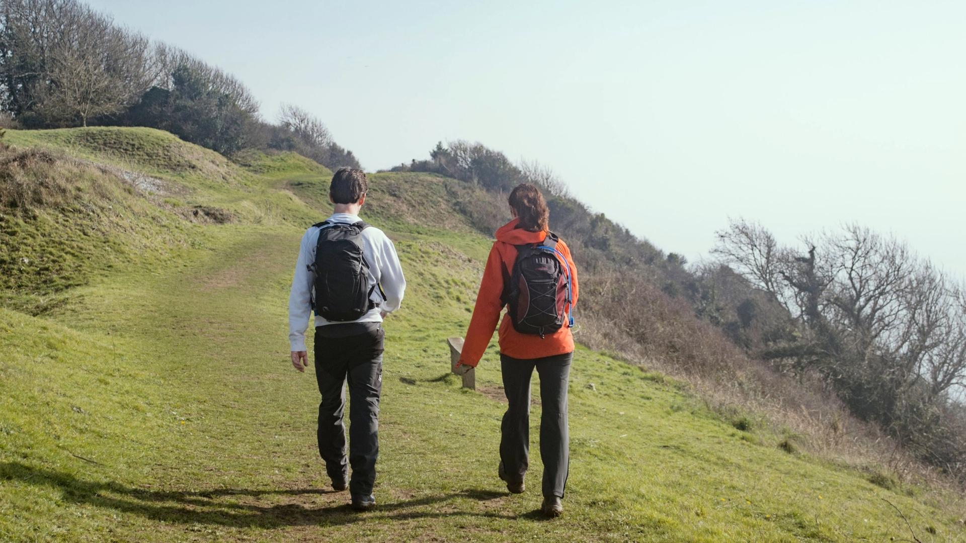 Two people with backpacks walk up a grassy hill on a sunny day.