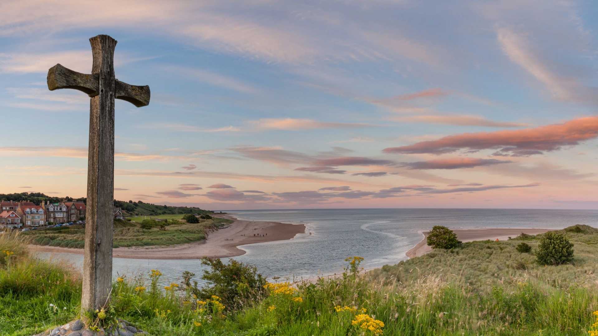 A wooden cross stands in the foreground overlooking a coastal landscape with a river meeting the sea under a colourful sunset sky.
