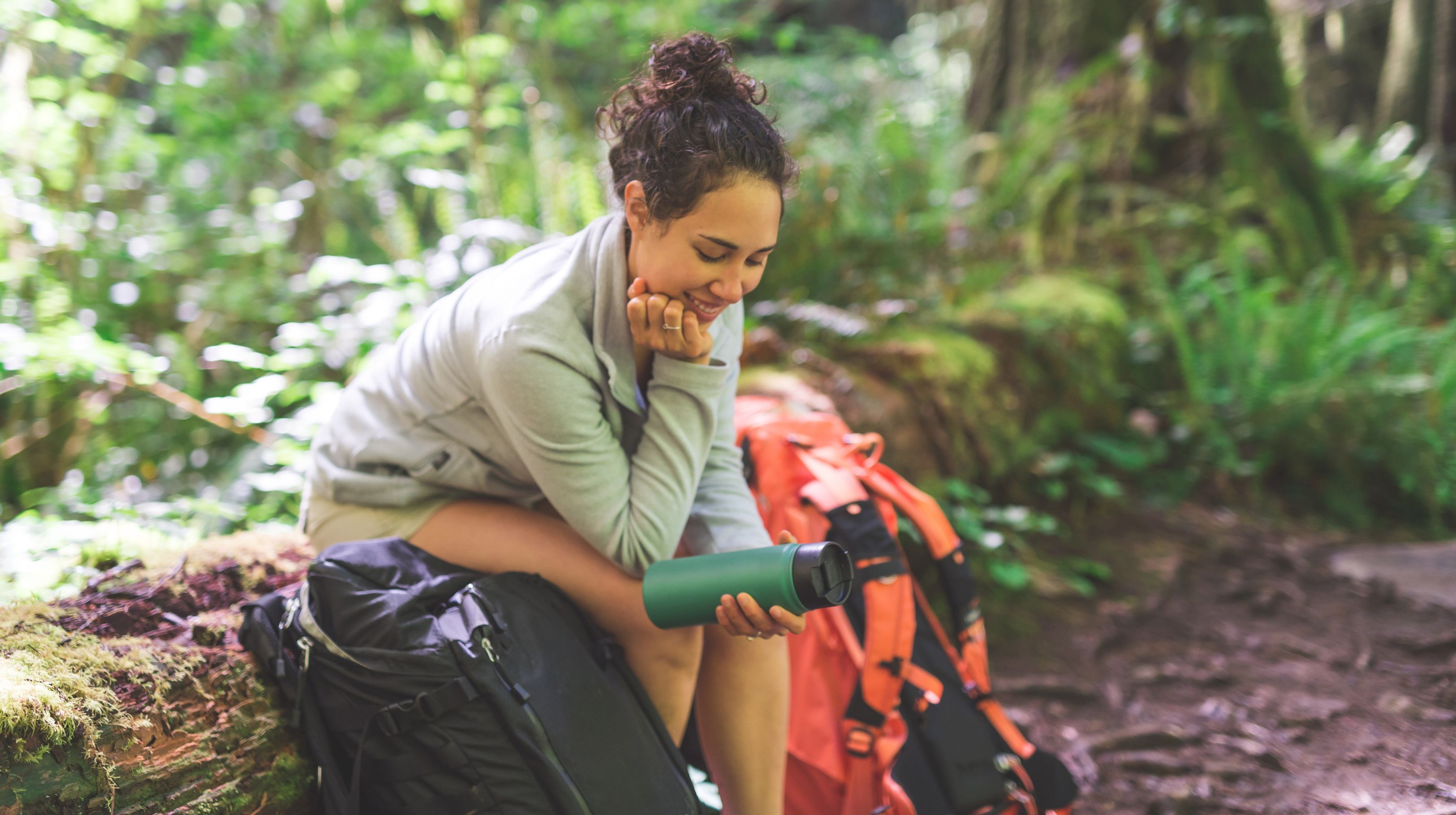 A person sitting on a log in a forest, holding a green water bottle, with backpacks nearby.