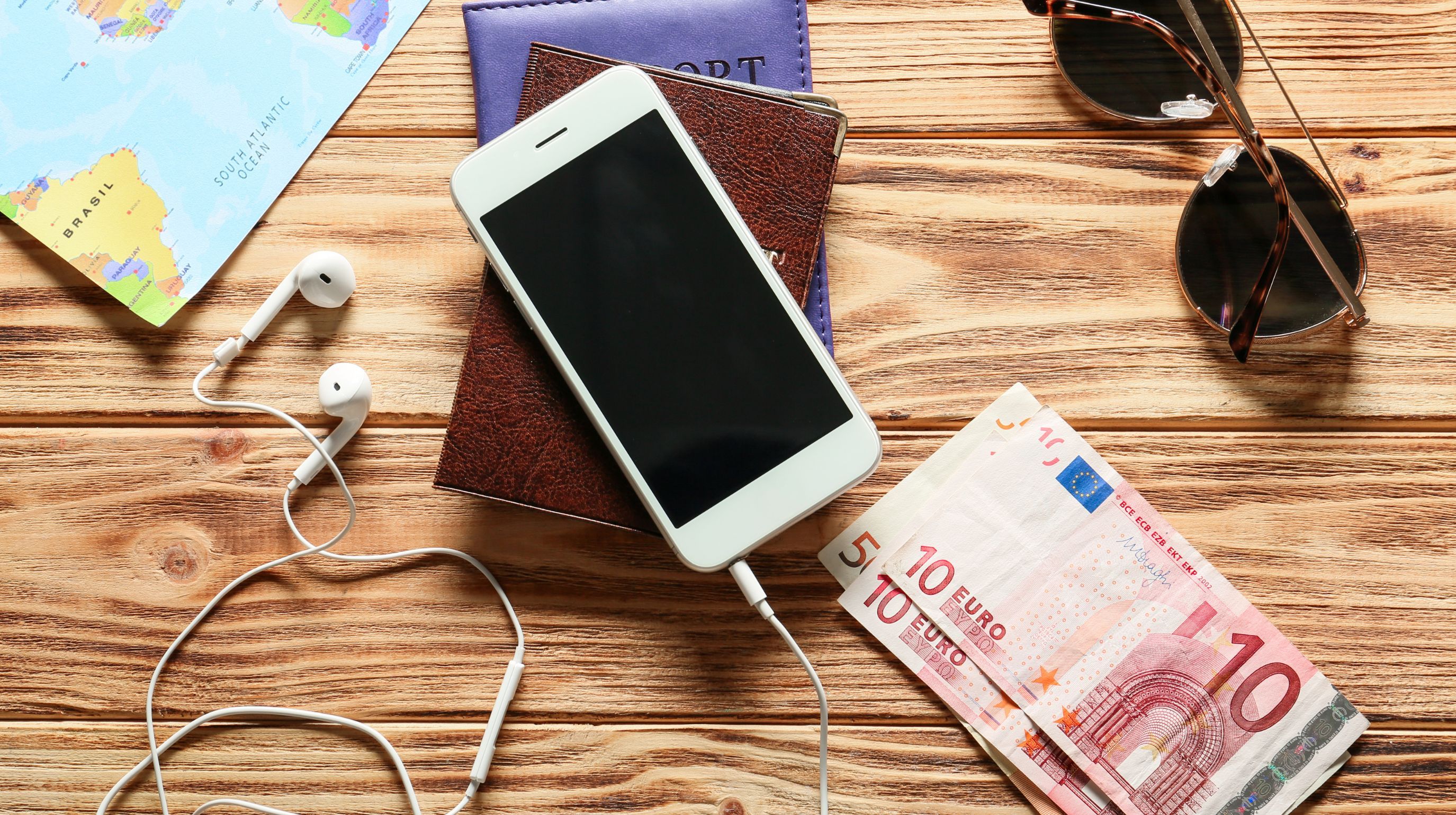 A collection of travel essentials including a map, passports, smartphone, earphones, sunglasses, and euro banknotes laid out on a wooden surface.