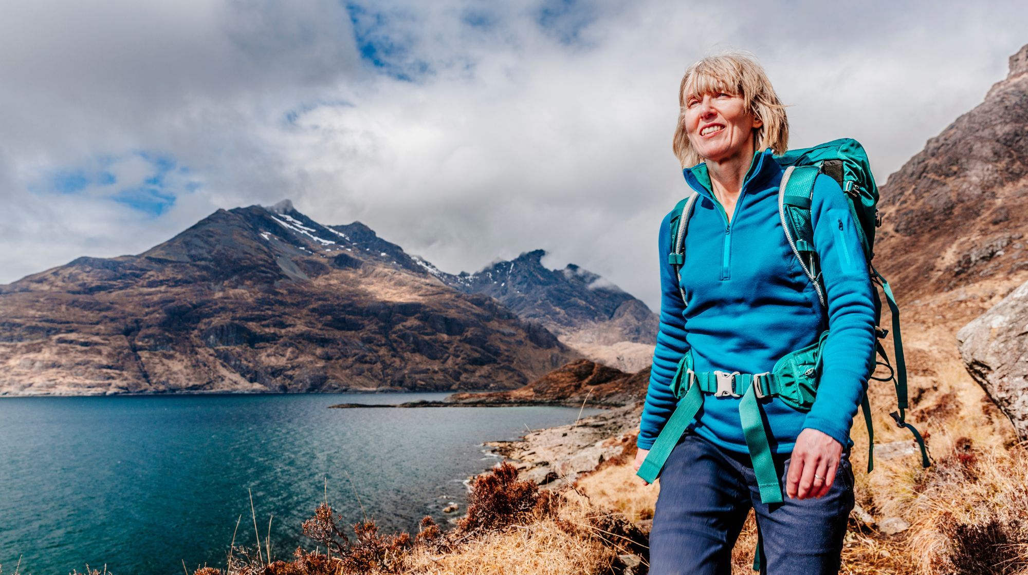 A person in a blue fleece and backpack stands on a trail overlooking a lake and mountains.