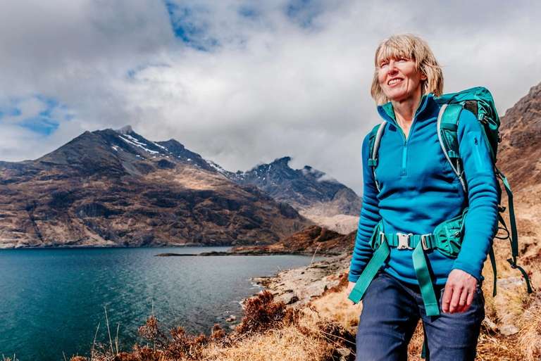 A person in a blue fleece and backpack stands on a trail overlooking a lake and mountains.