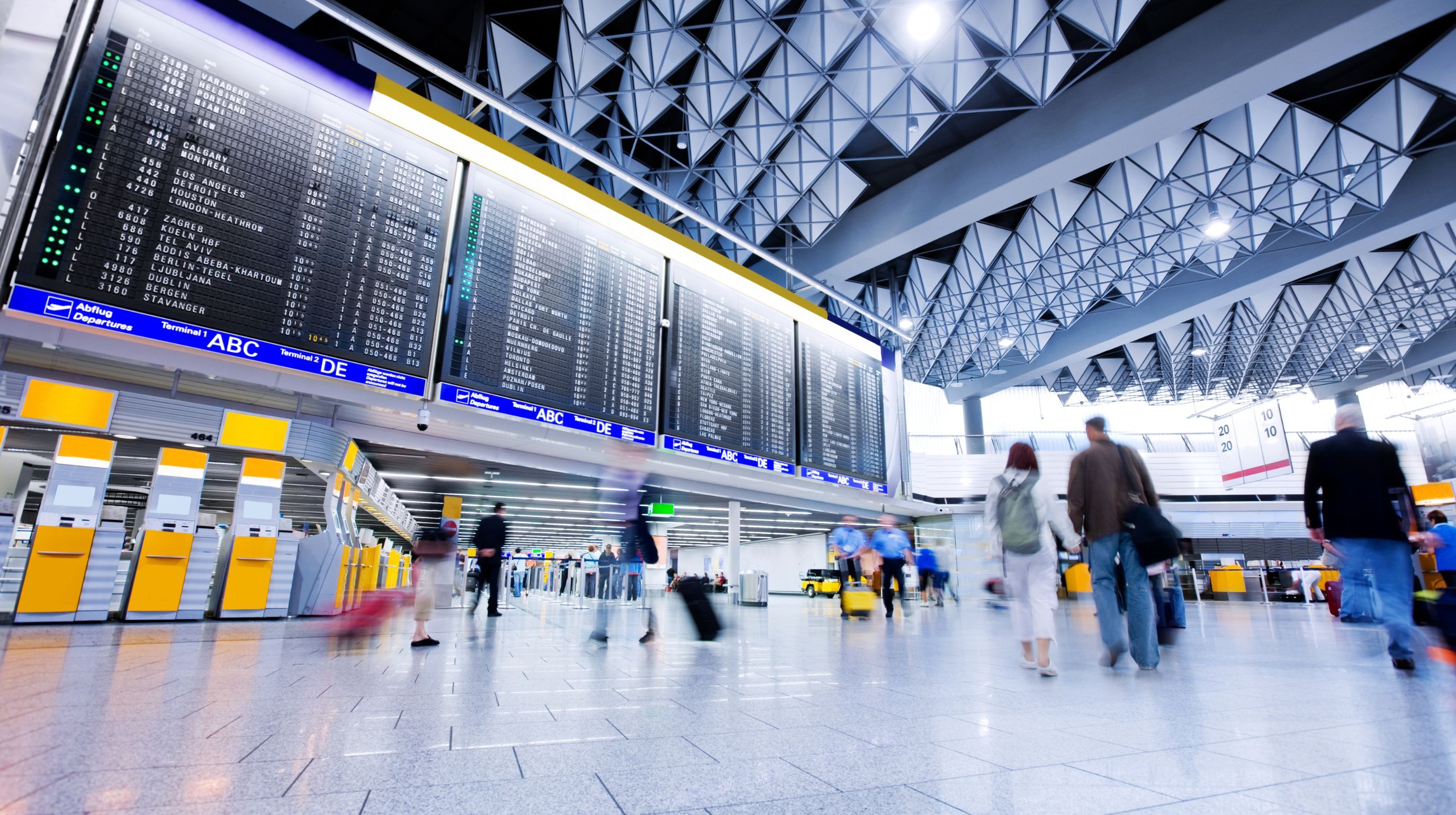 A large departure board displays flight information in a busy airport terminal with people walking and pulling luggage.