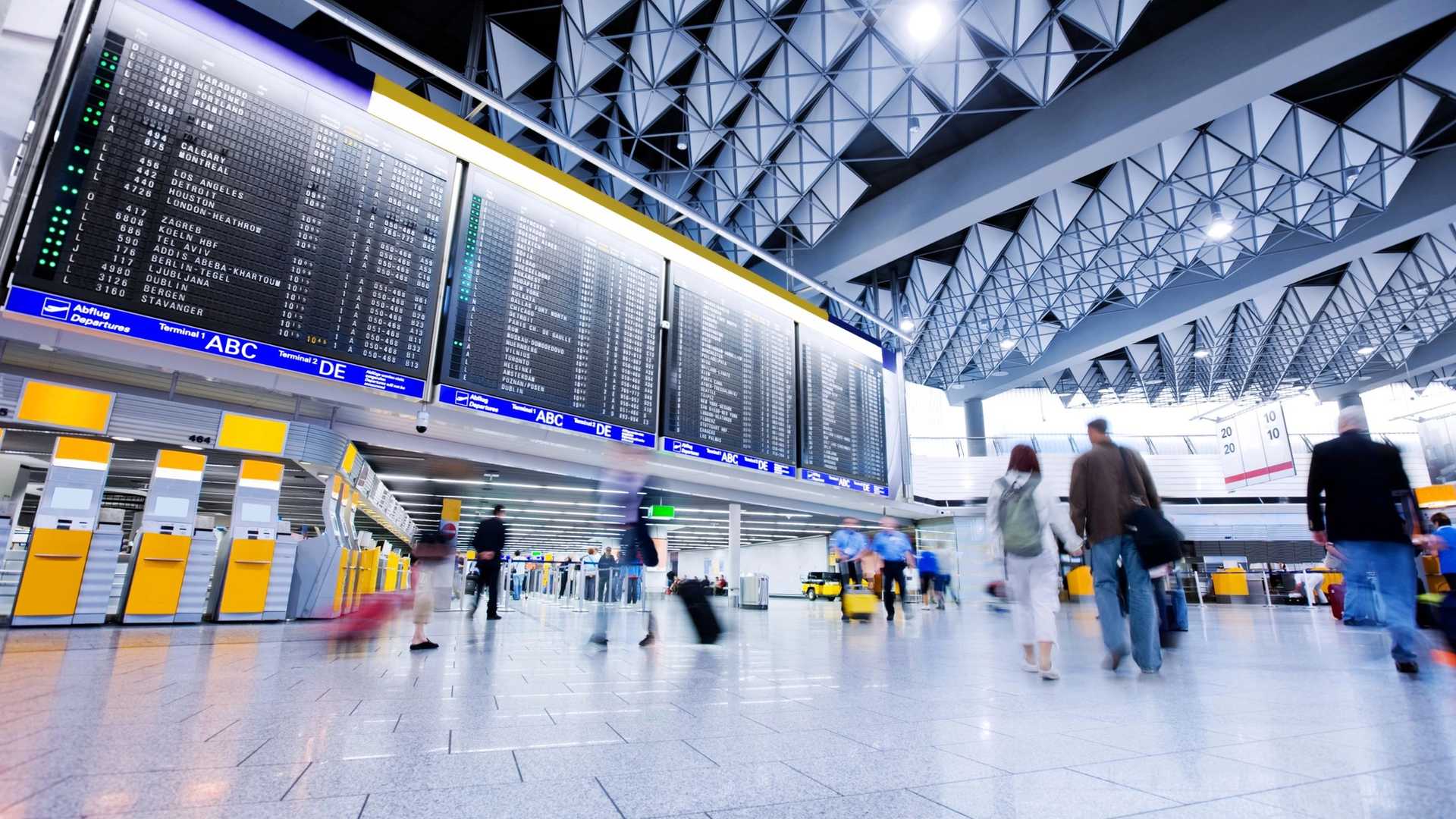A large departure board displays flight information in a busy airport terminal with people walking and pulling luggage.