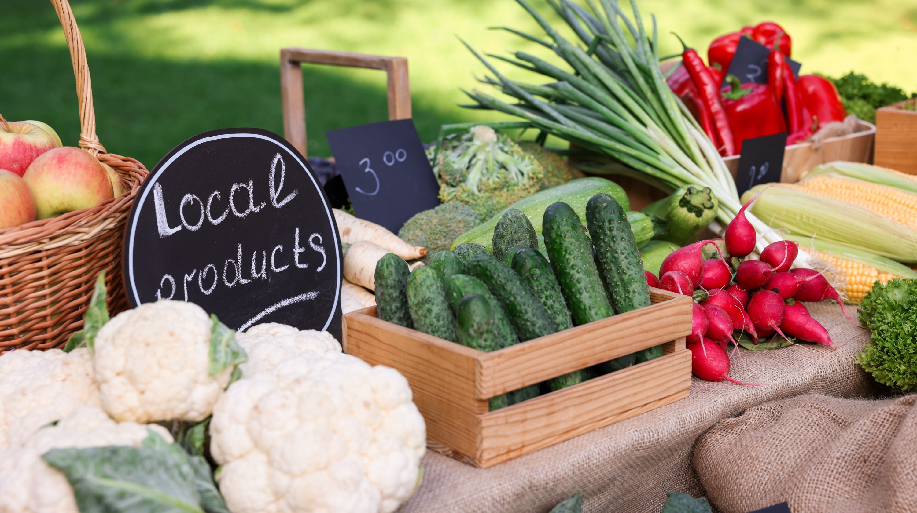 A wooden crate of cucumbers sits on a table displaying a variety of fresh produce, including cauliflower, apples, radishes, corn, and peppers, with a sign that reads "Local products."