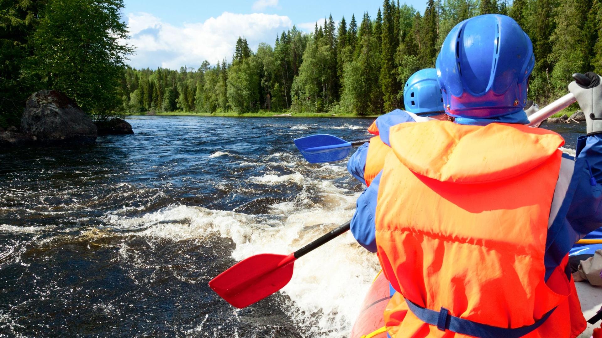 Two people in orange life vests and blue helmets are white water rafting on a river, holding red and blue paddles.