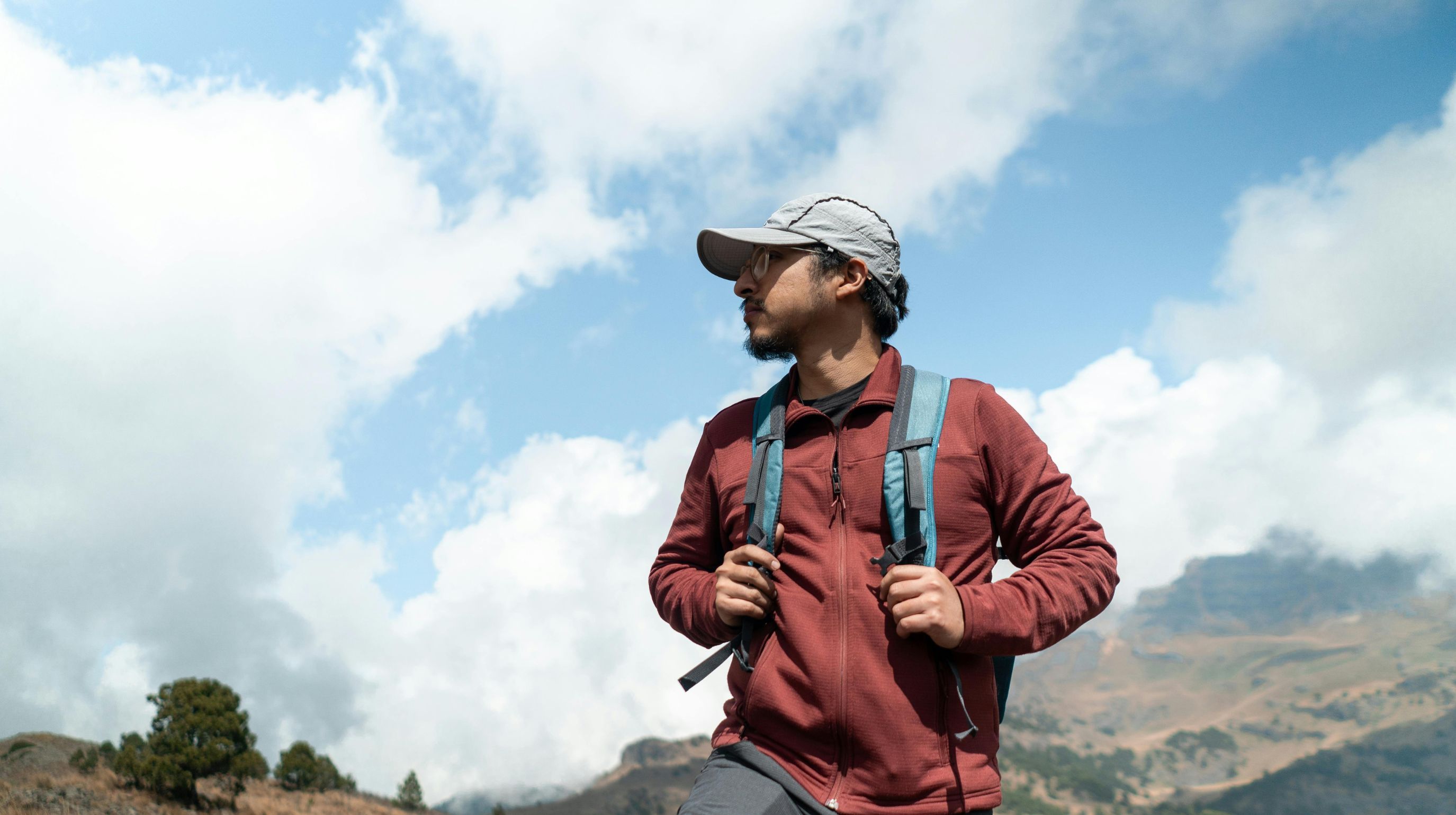 A person wearing a maroon jacket and a cap stands on a mountain with a backpack, looking off into the distance under a cloudy sky.