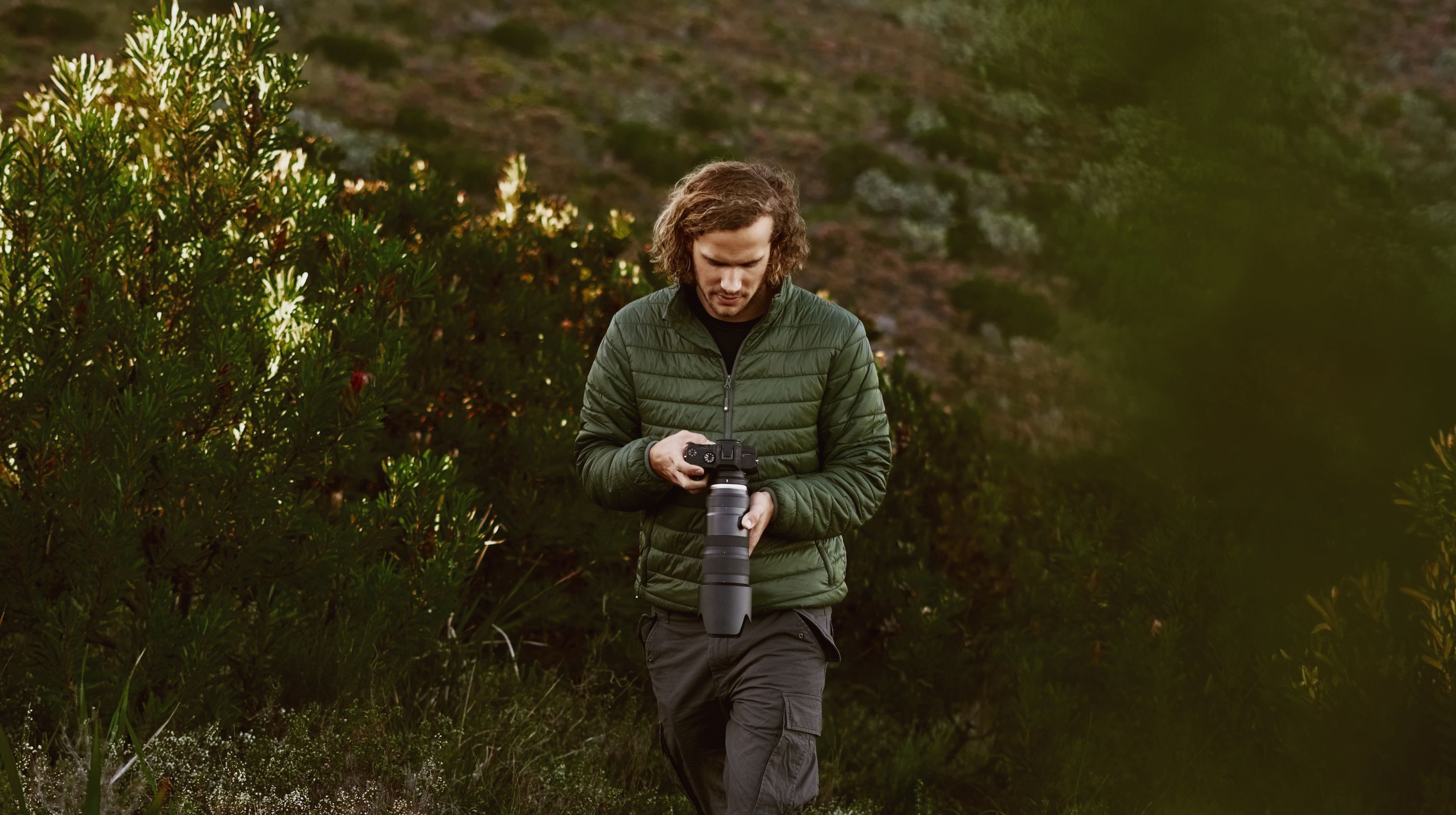 A person with curly hair wearing a green puffer jacket and cargo pants holds a camera with a long lens in a natural setting with green foliage.