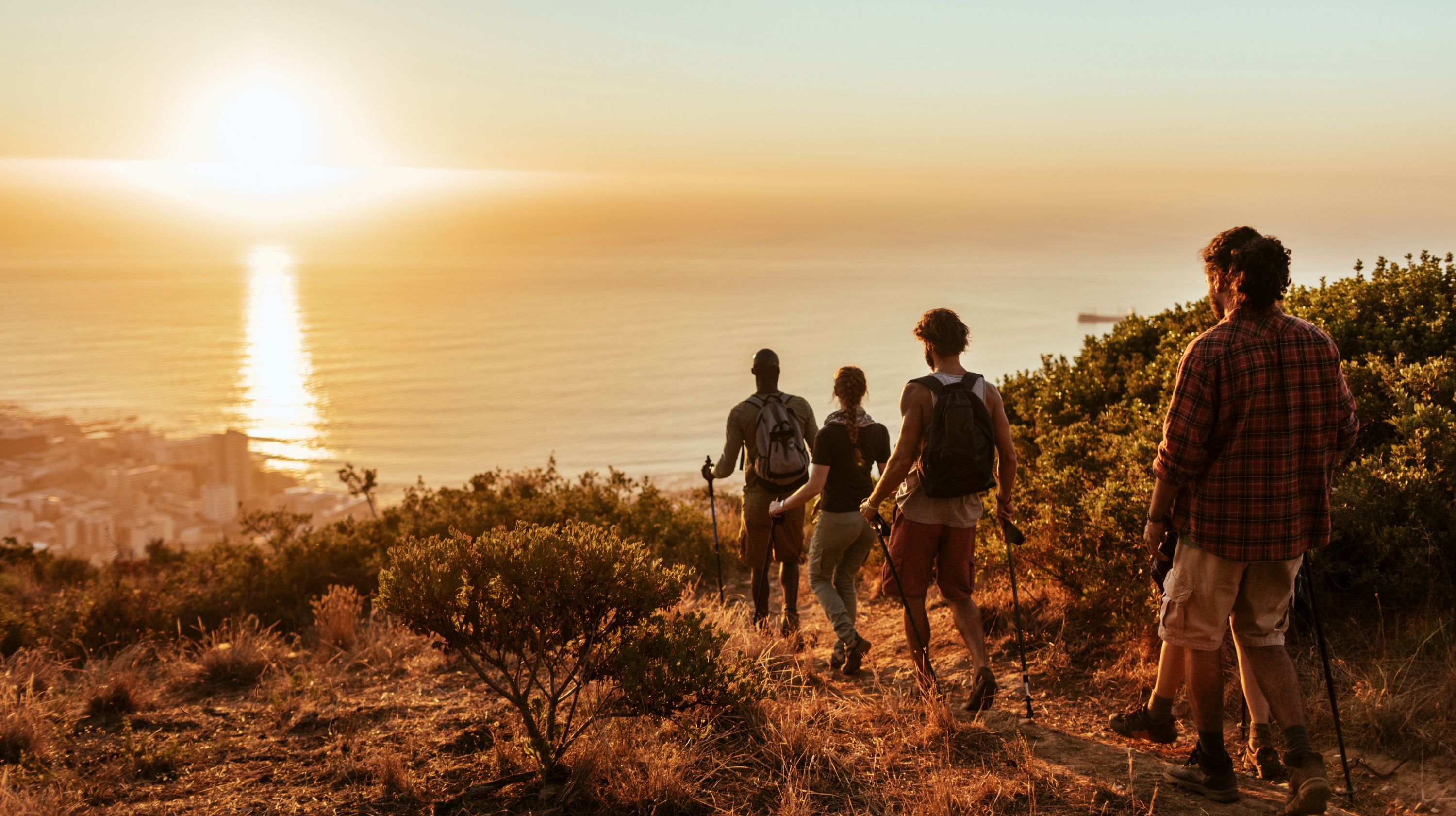 A group of hikers walk along a ridge overlooking the ocean at sunset, with a city visible in the distance.