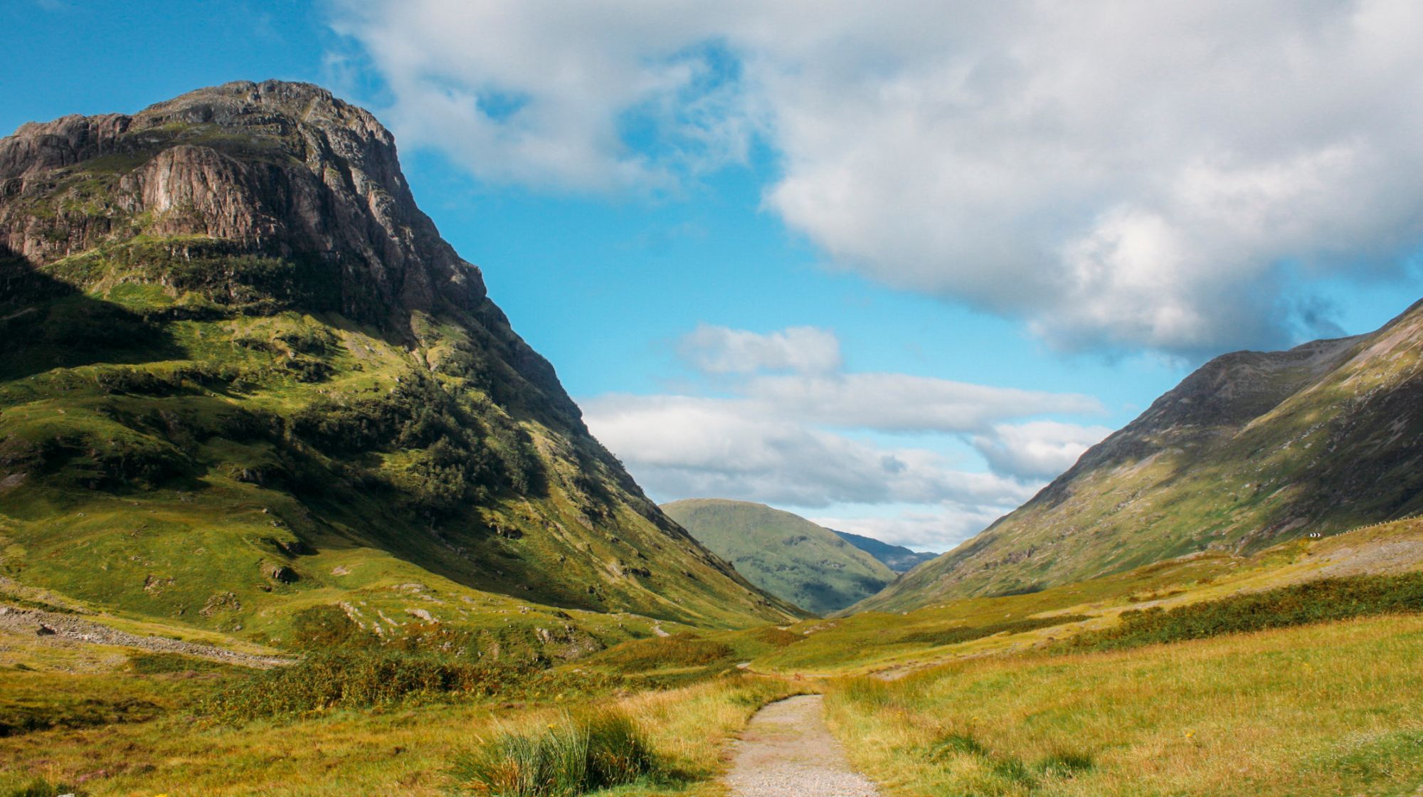 A scenic view of a valley with rolling green hills and a path leading into the distance under a partly cloudy sky.