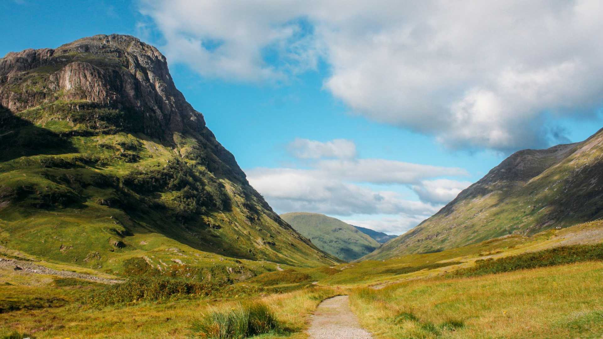A scenic view of a valley with rolling green hills and a path leading into the distance under a partly cloudy sky.