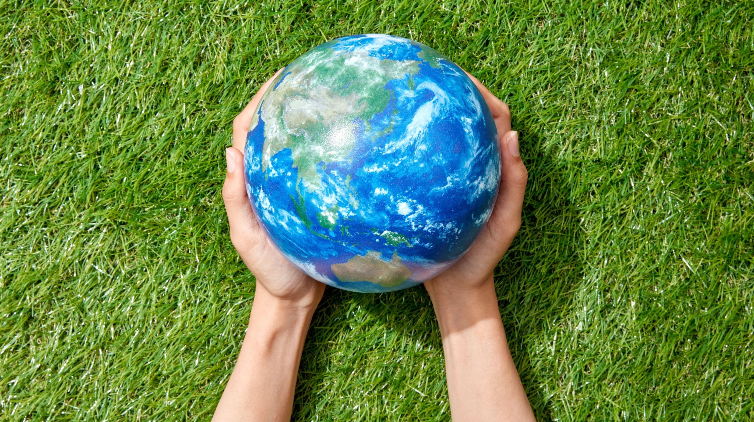 Two hands cradle a model of the Earth against a backdrop of green grass.