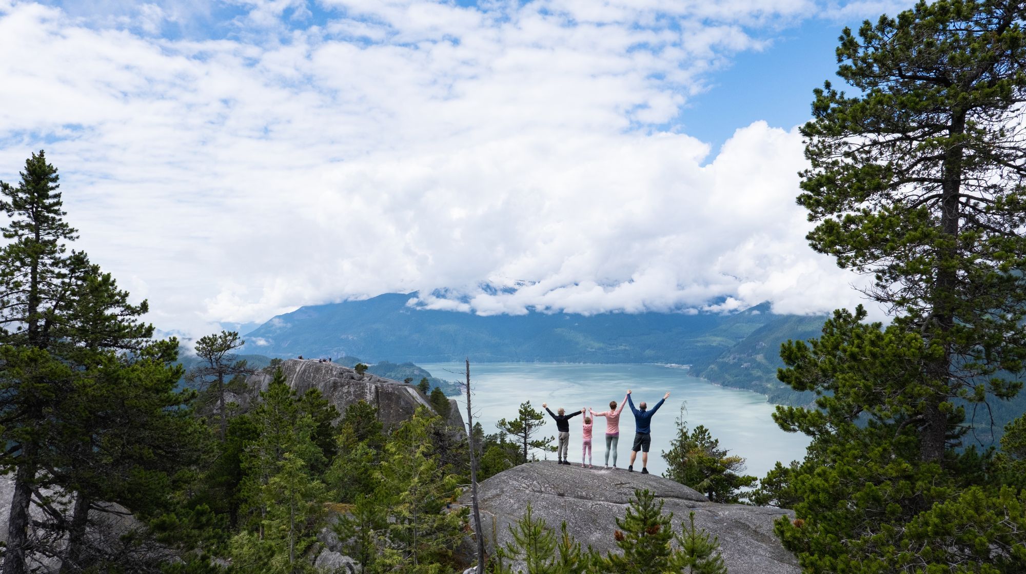 A group of people stand on a rocky outcrop with their arms raised, overlooking a vast body of water surrounded by mountains under a cloudy sky.