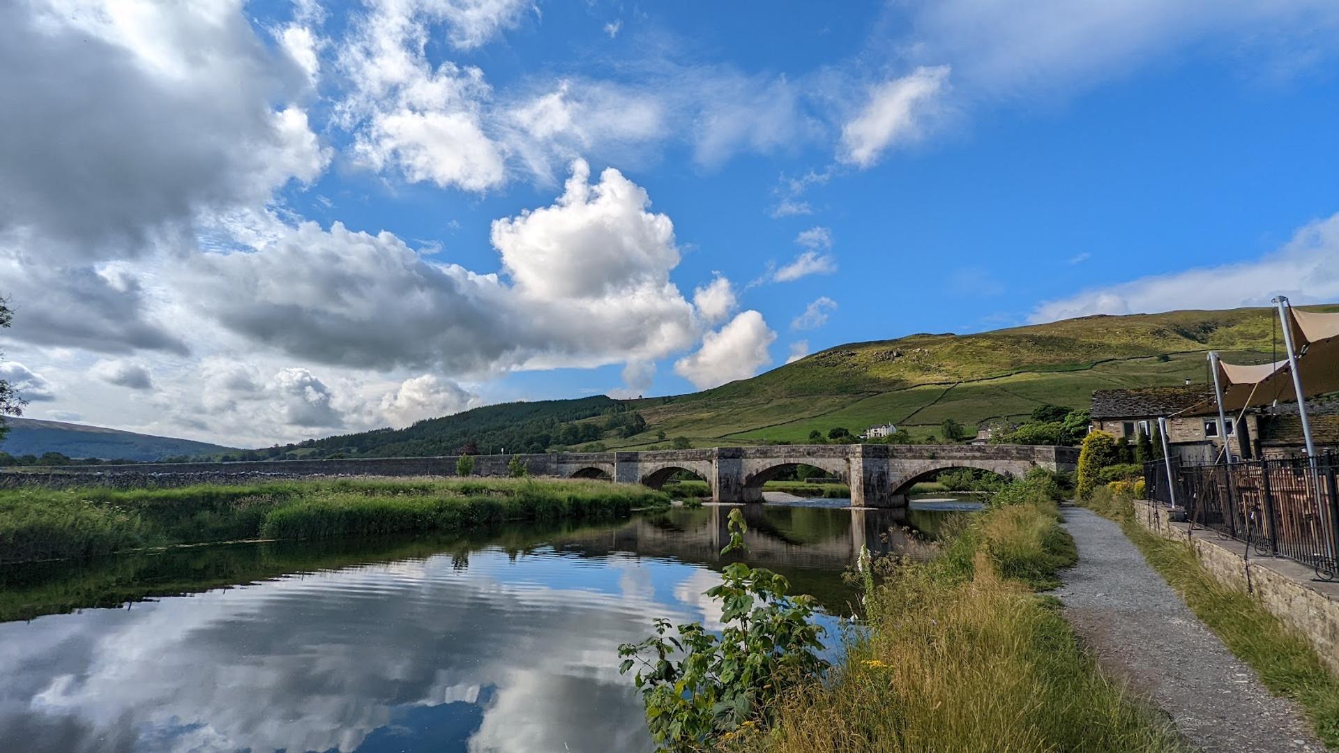 Riverside path and historic stone bridge at Burnsall on the Dales Way, surrounded by the rolling landscapes of the Yorkshire Dales.