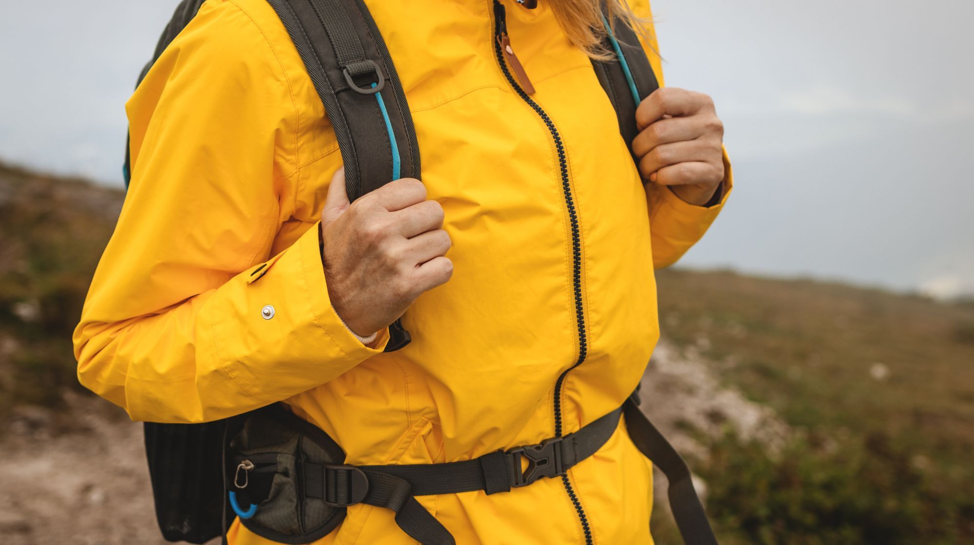 A person wearing a bright yellow raincoat and backpack stands on a trail with blurred natural scenery in the background.