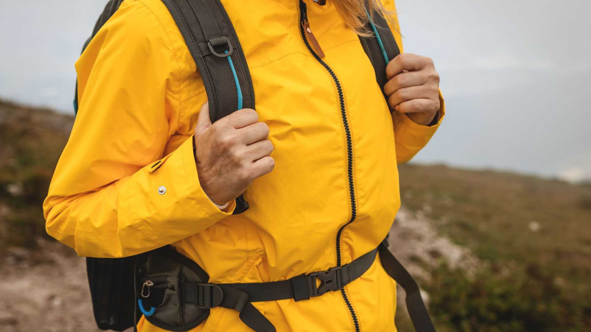 A person wearing a bright yellow raincoat and backpack stands on a trail with blurred natural scenery in the background.