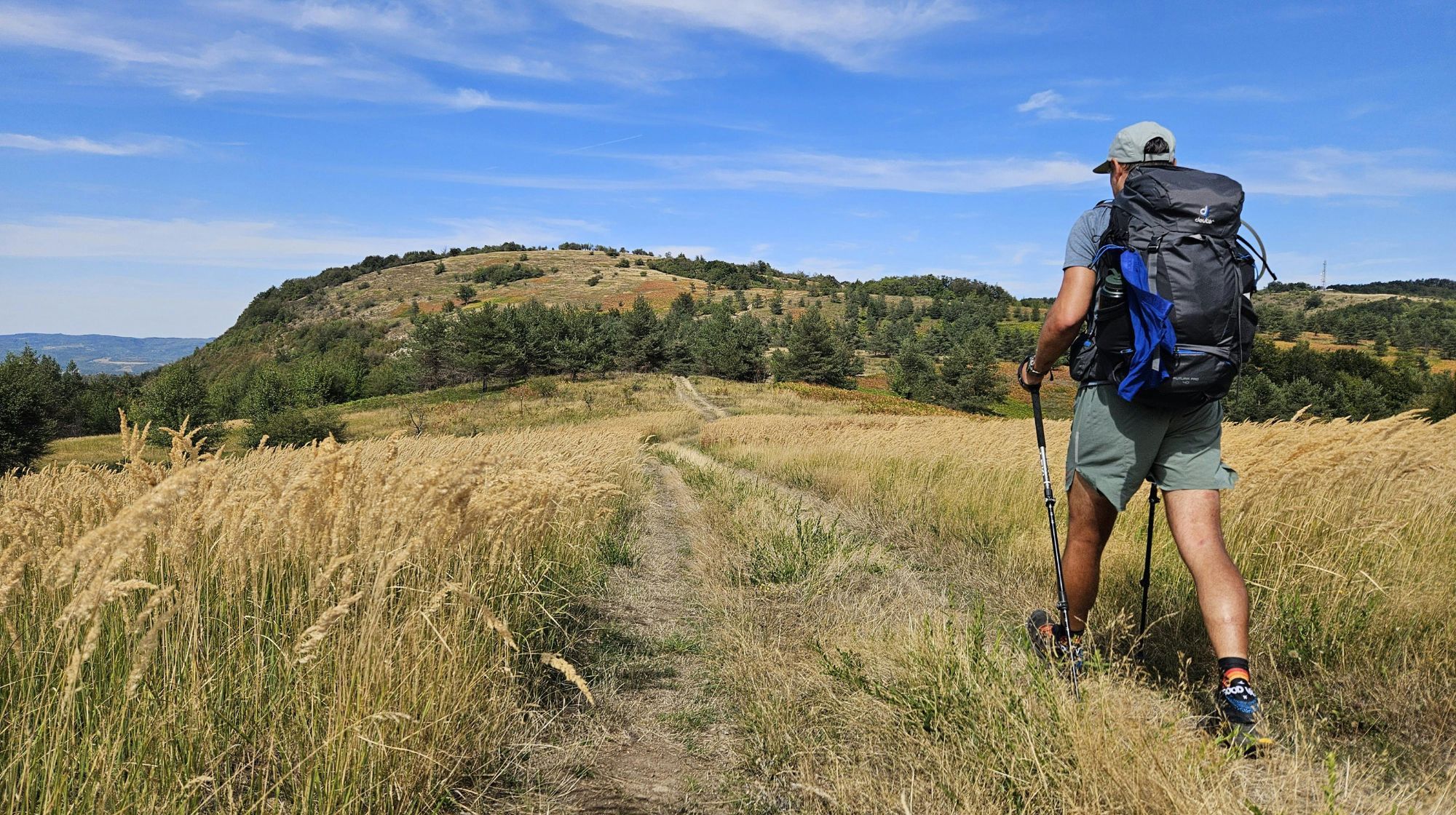 A person with a large backpack hikes along a path through a field of dry grass under a blue sky with scattered clouds. A gentle hill covered in green trees and shrubs is visible in the background.